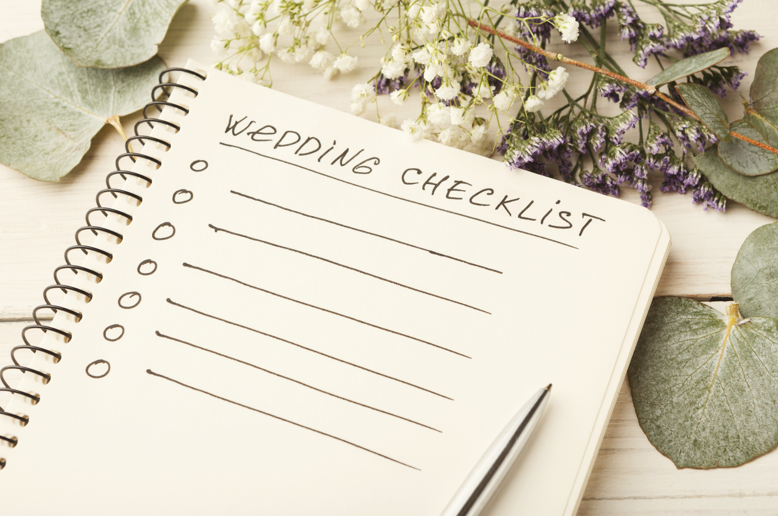 A checklist titled 'Wedding Checklist' on a spiral notebook with a black pen, surrounded by white and purple flowers and green leaves on a light wooden surface.