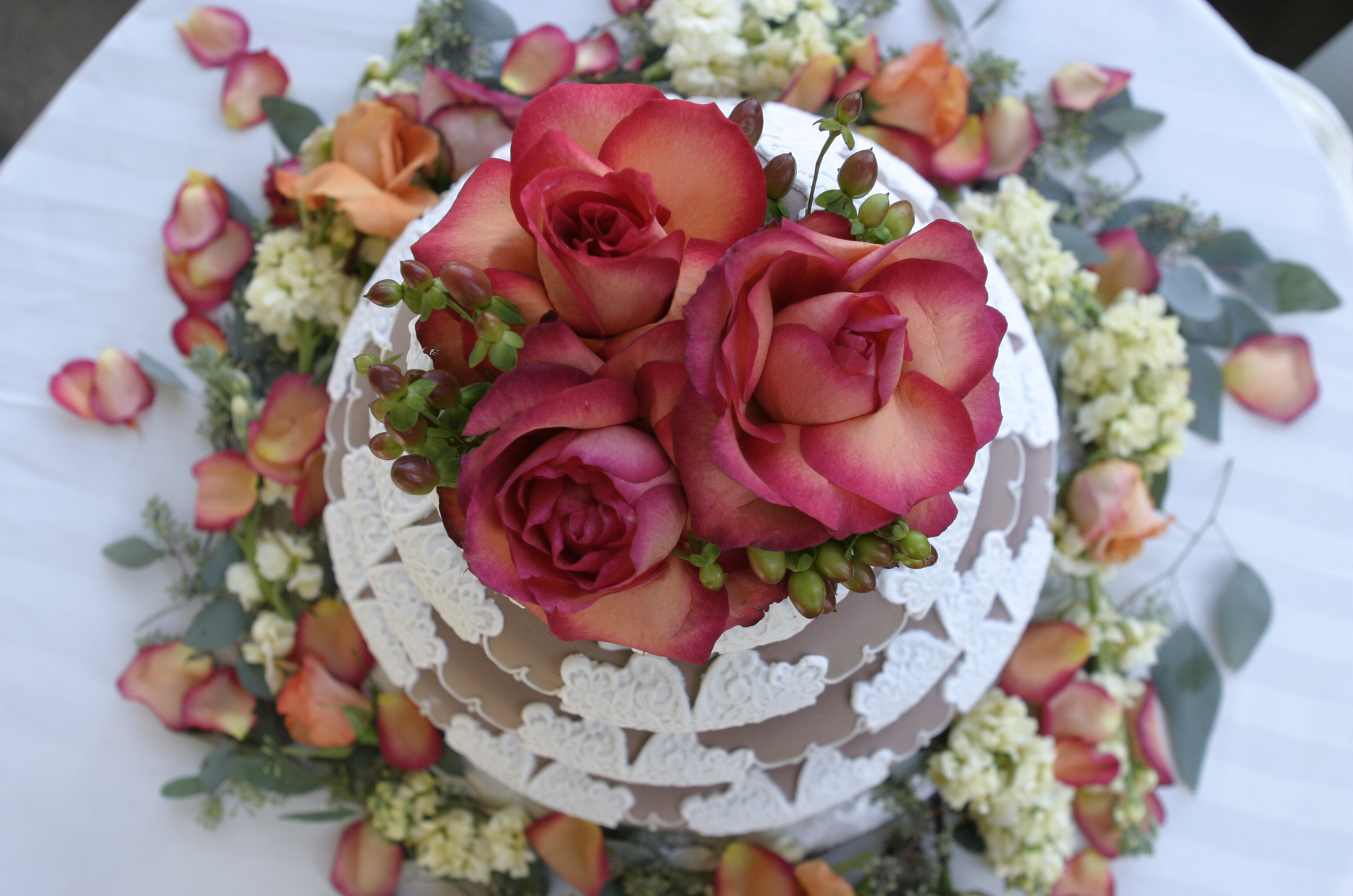 A wedding cake decorated with a bouquet of pink and orange roses on top, surrounded by rose petals and additional small flowers and greenery.