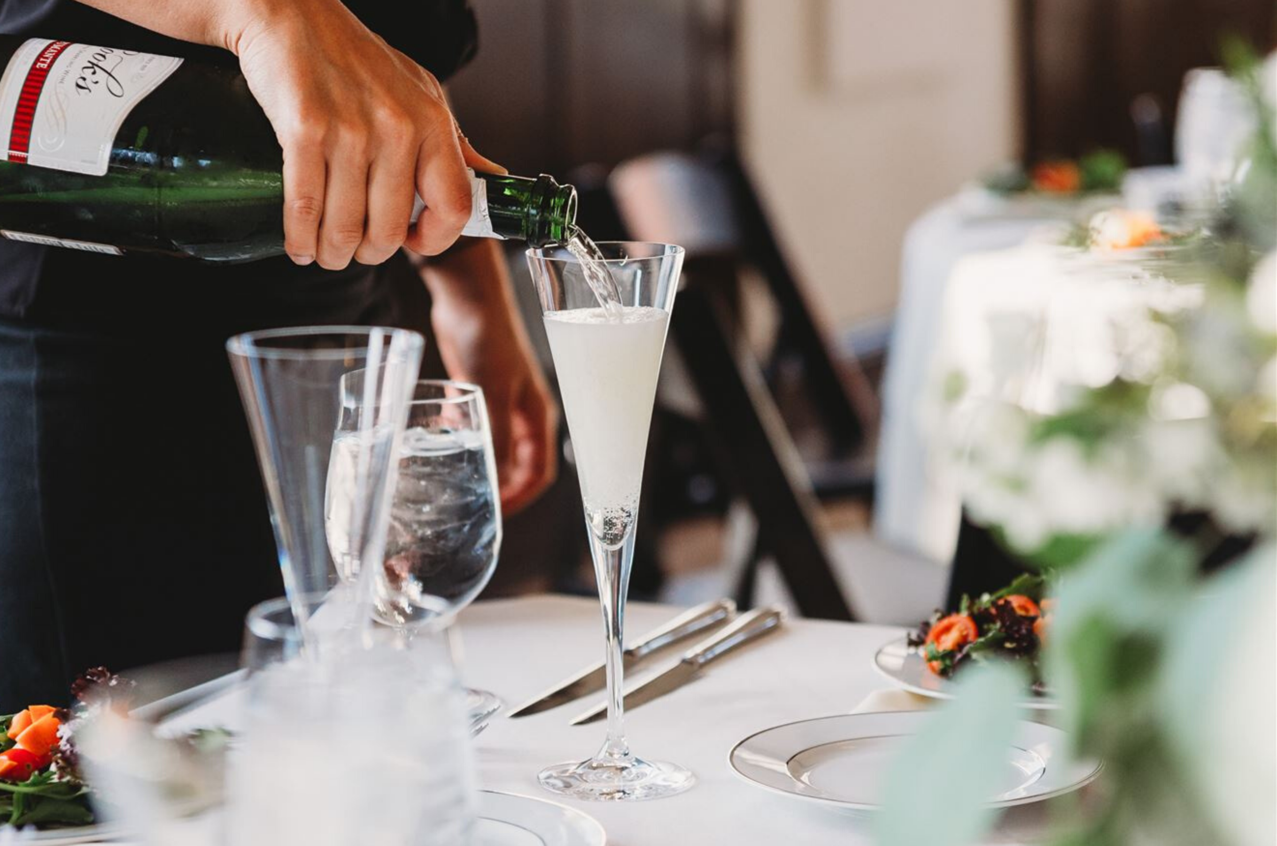 Someone pouring a clear sparkling beverage into a tall champagne flute on a formal dining table.