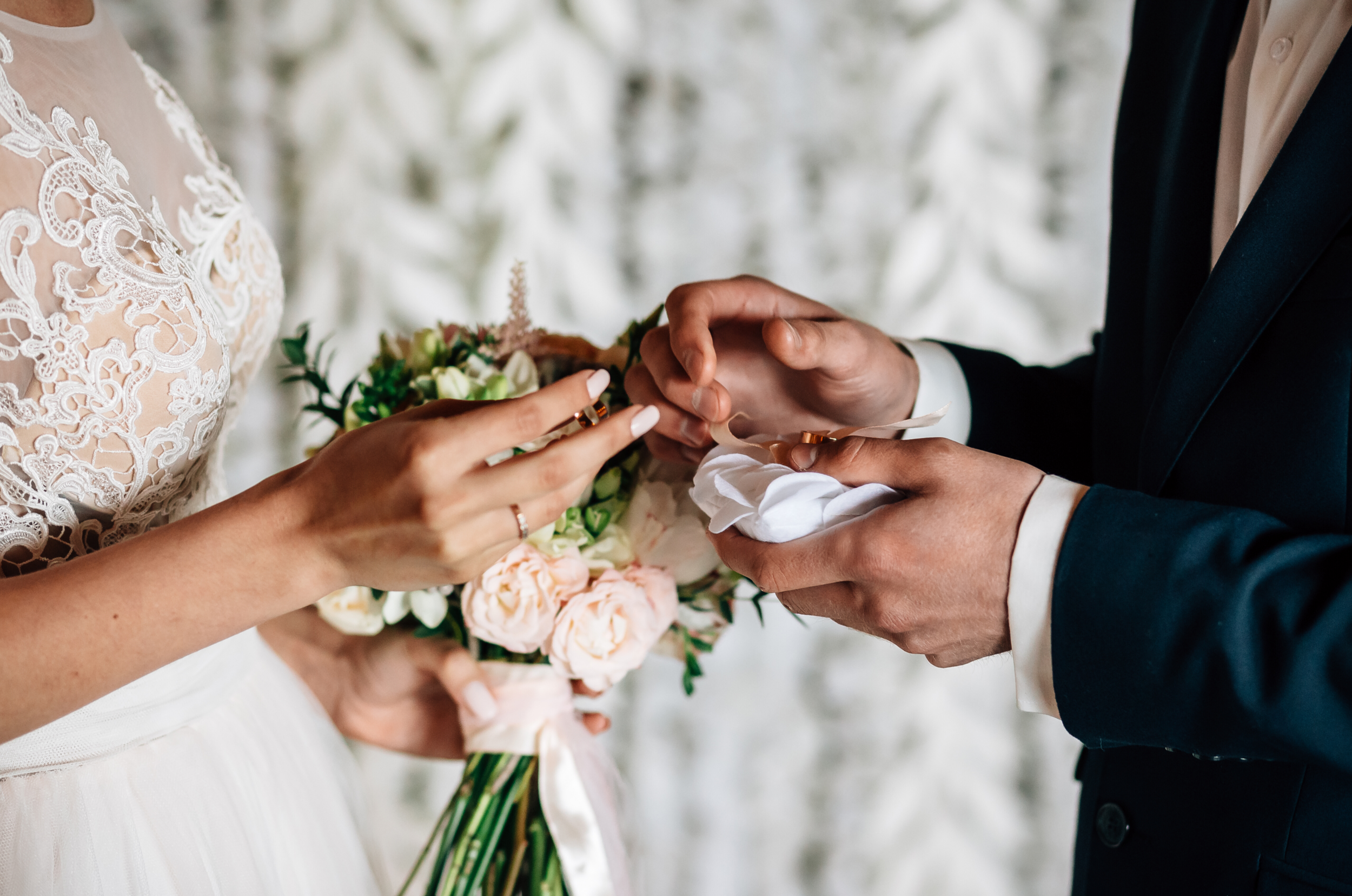 A bride and groom exchange rings during their wedding ceremony. The bride wears a lace dress and holds a bouquet of pink and white flowers, while the groom wears a black suit.