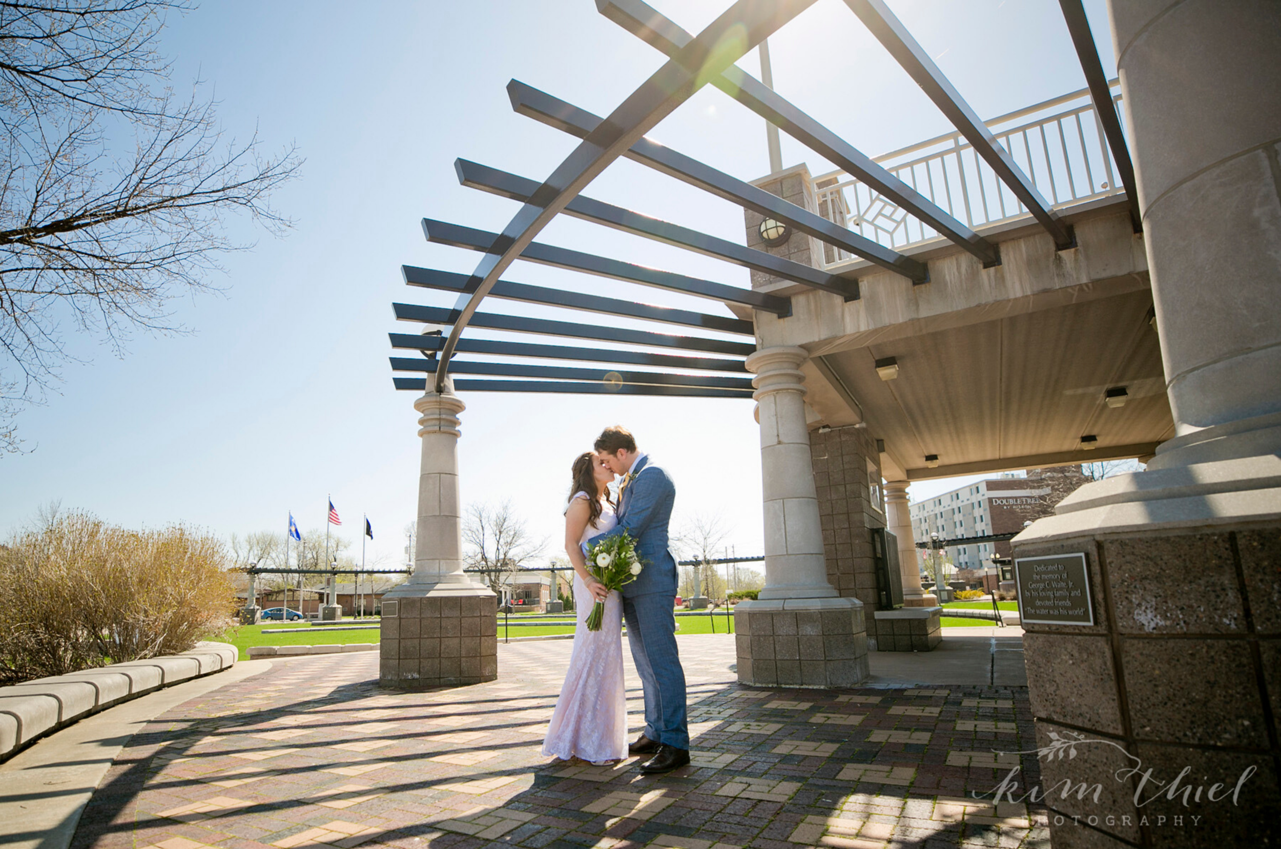 A bride and groom standing under a pergola, kissing, with the bride holding a bouquet, in a park on a sunny day.