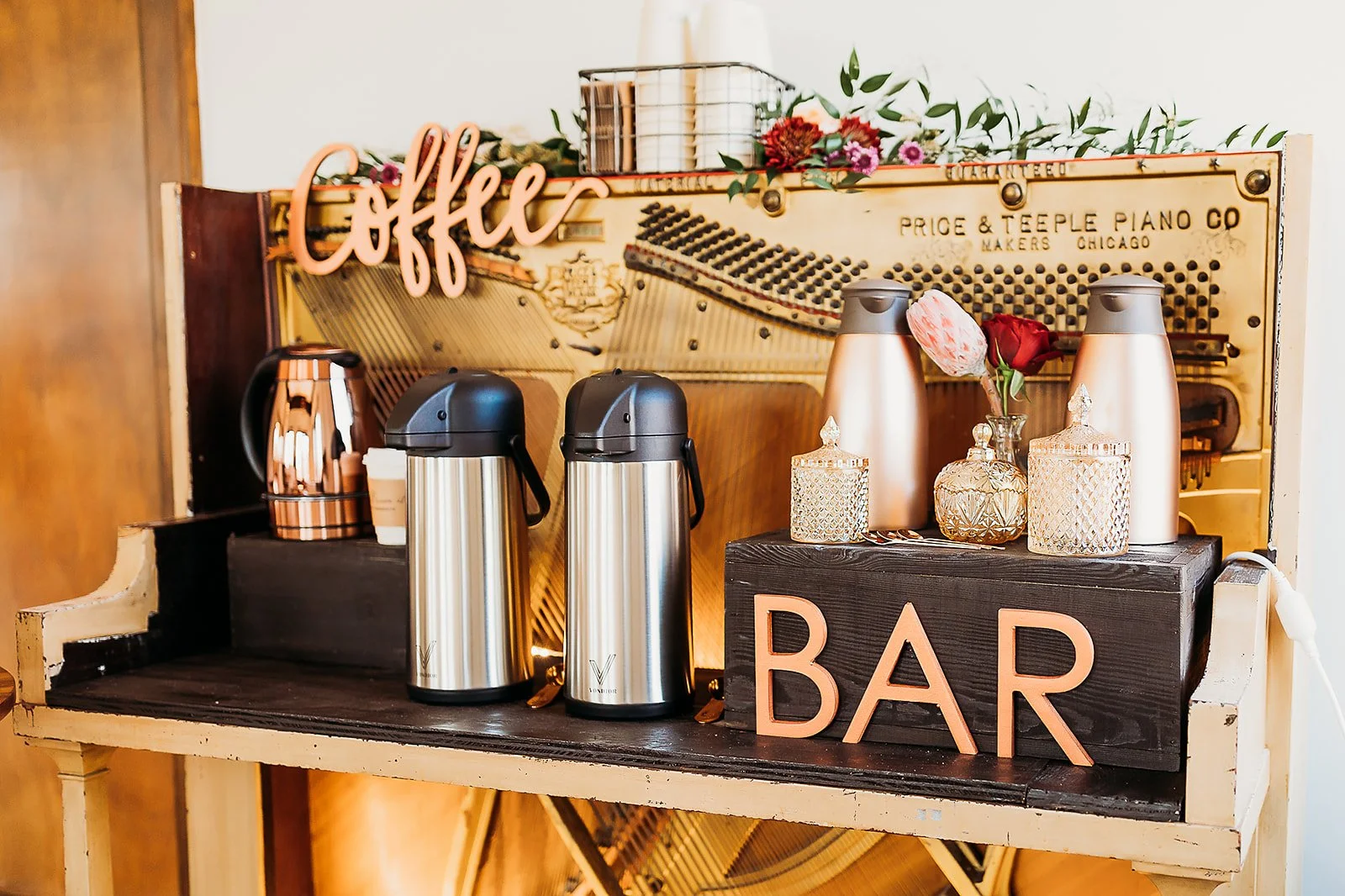 Coffee bar setup with stainless steel thermoses, decorative jars, pink floral sign, and a black wooden sign that says 'Bar' at the front, with a vintage piano and floral decoration in the background.