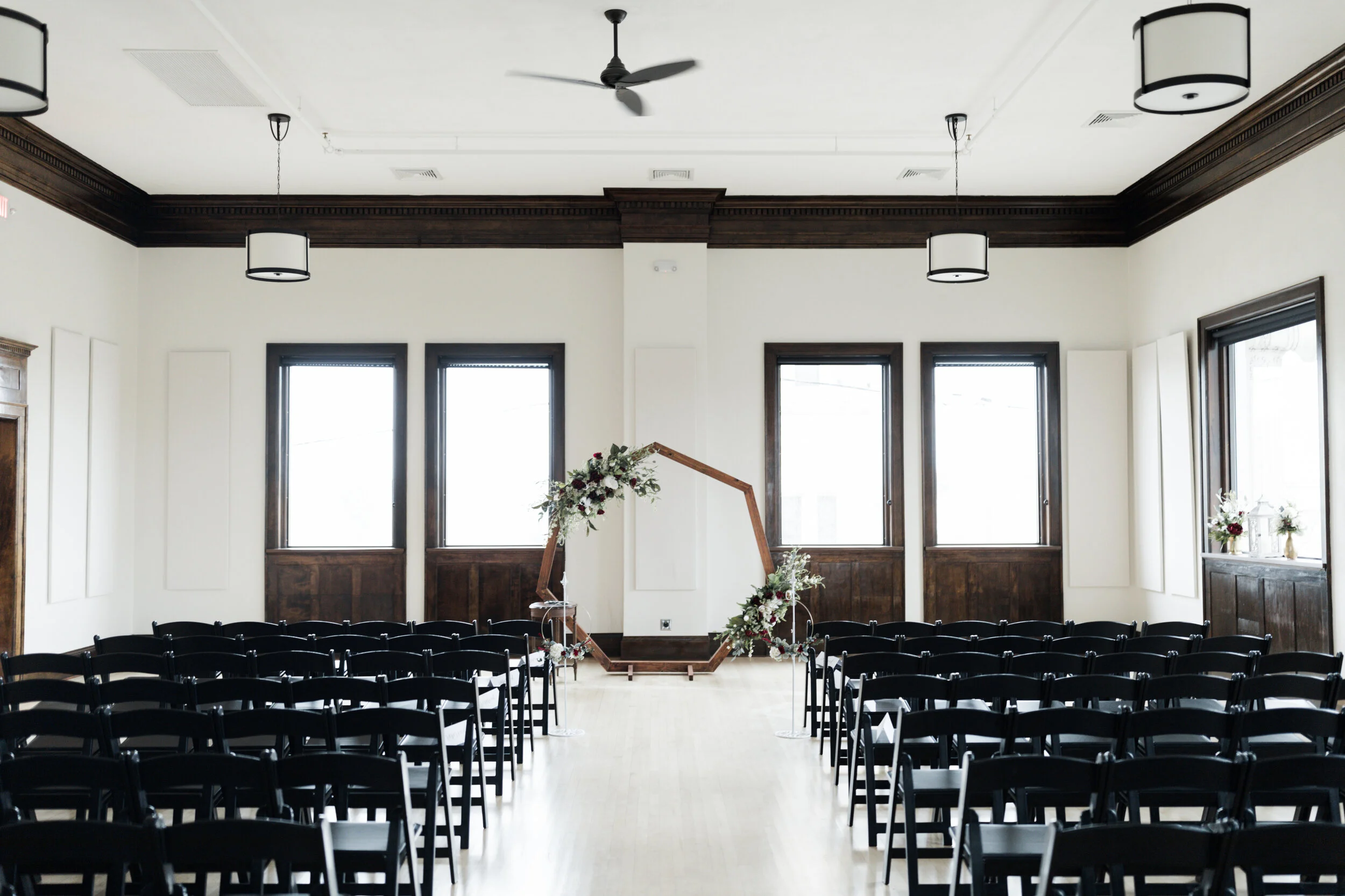 Empty indoor wedding ceremony space with rows of black chairs facing a floral geometric arch at the front, dark wood trim around windows, and minimal decor.