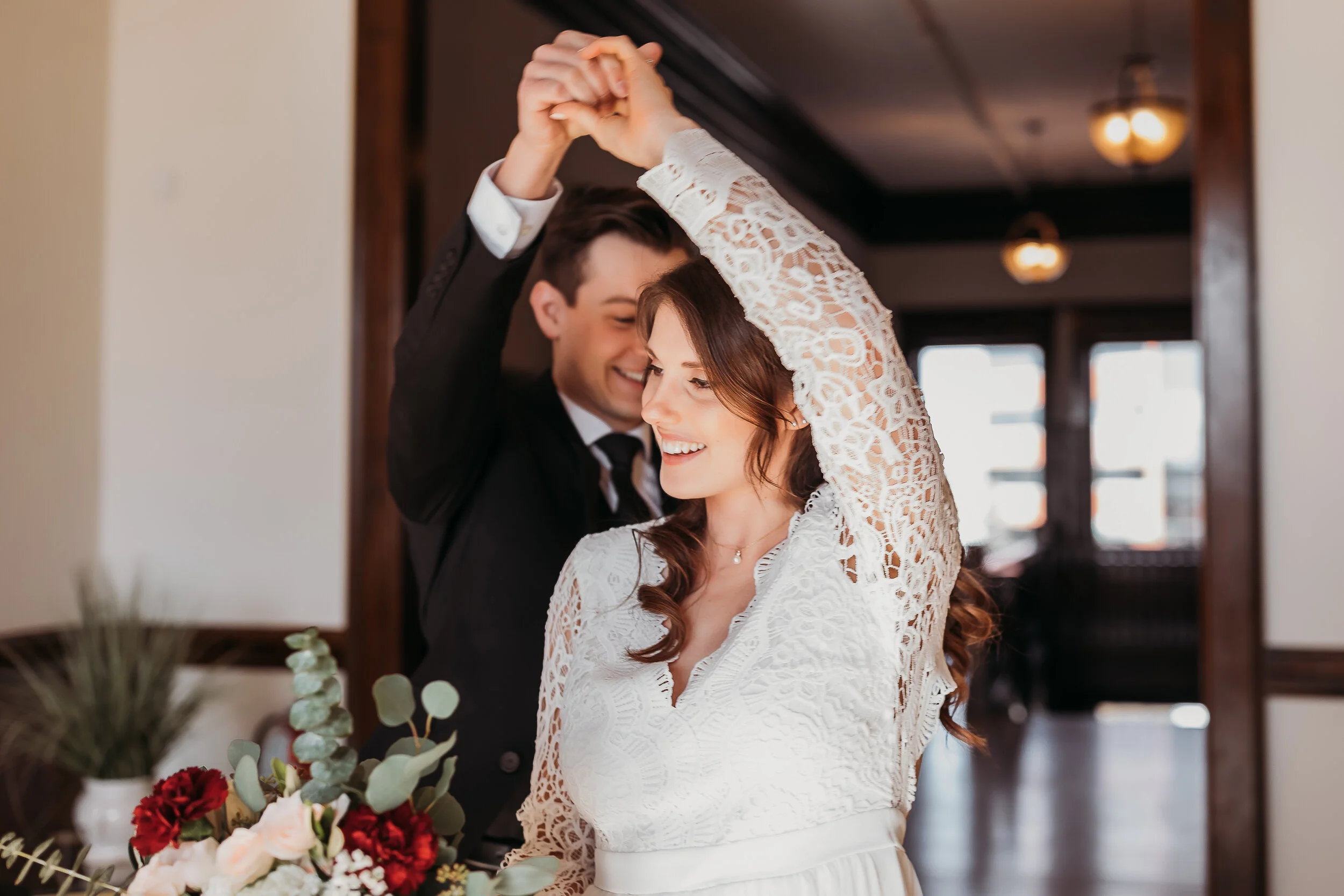 A couple dancing indoors, smiling, with a bouquet of flowers on the table in front of them.