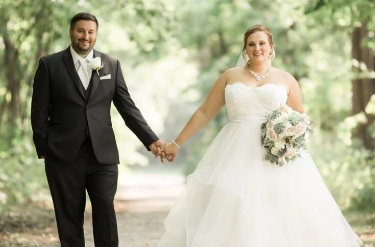 A bride and groom holding hands, outdoors in a forest setting, on their wedding day. The bride is wearing a white gown and holding a bouquet of flowers, and the groom is dressed in a black suit with a white shirt and tie.
