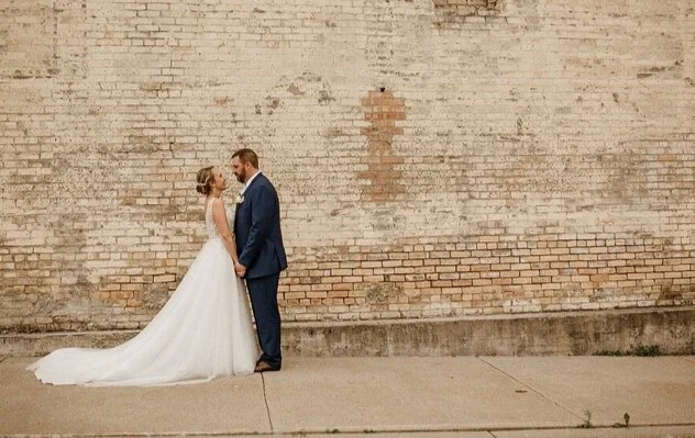 A bride and groom standing close together in front of a brick wall, holding hands and facing each other.