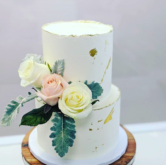 Two-tier white cake with gold leaf accents and floral decorations including white, pale pink, and yellow roses with green leaves, on a wooden cake stand.