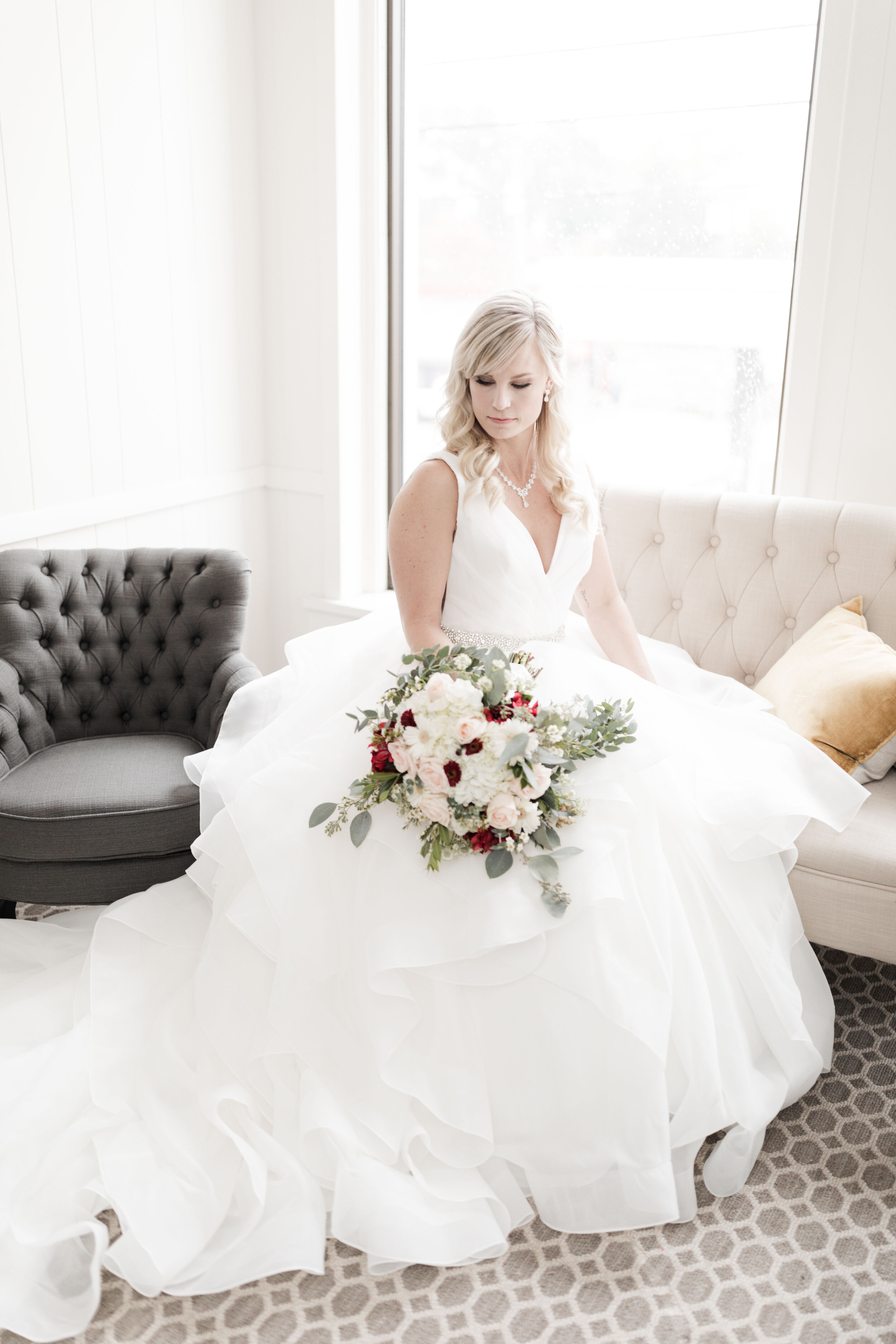 Bride sitting on a beige tufted sofa holding a bouquet of pink and red flowers with green foliage, wearing a white wedding dress, in a bright room with a large window.
