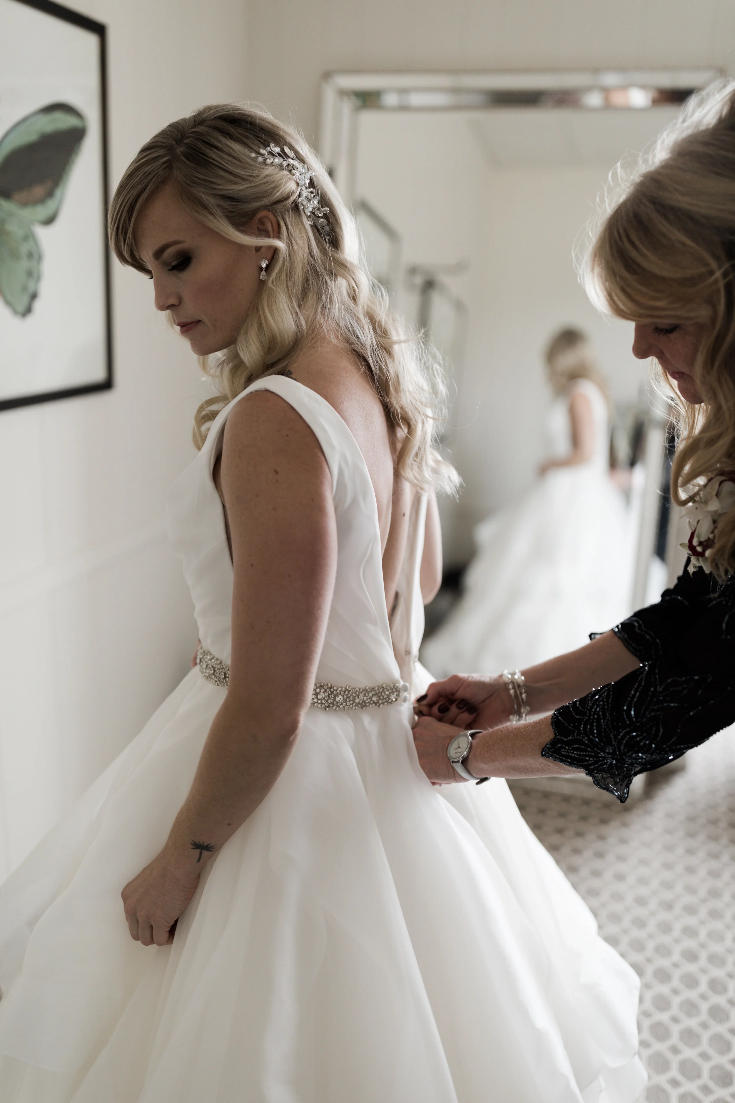 A bride in a white wedding gown with a bejeweled belt is being helped by a woman into her dress, with the bride also visible in a mirror reflection.