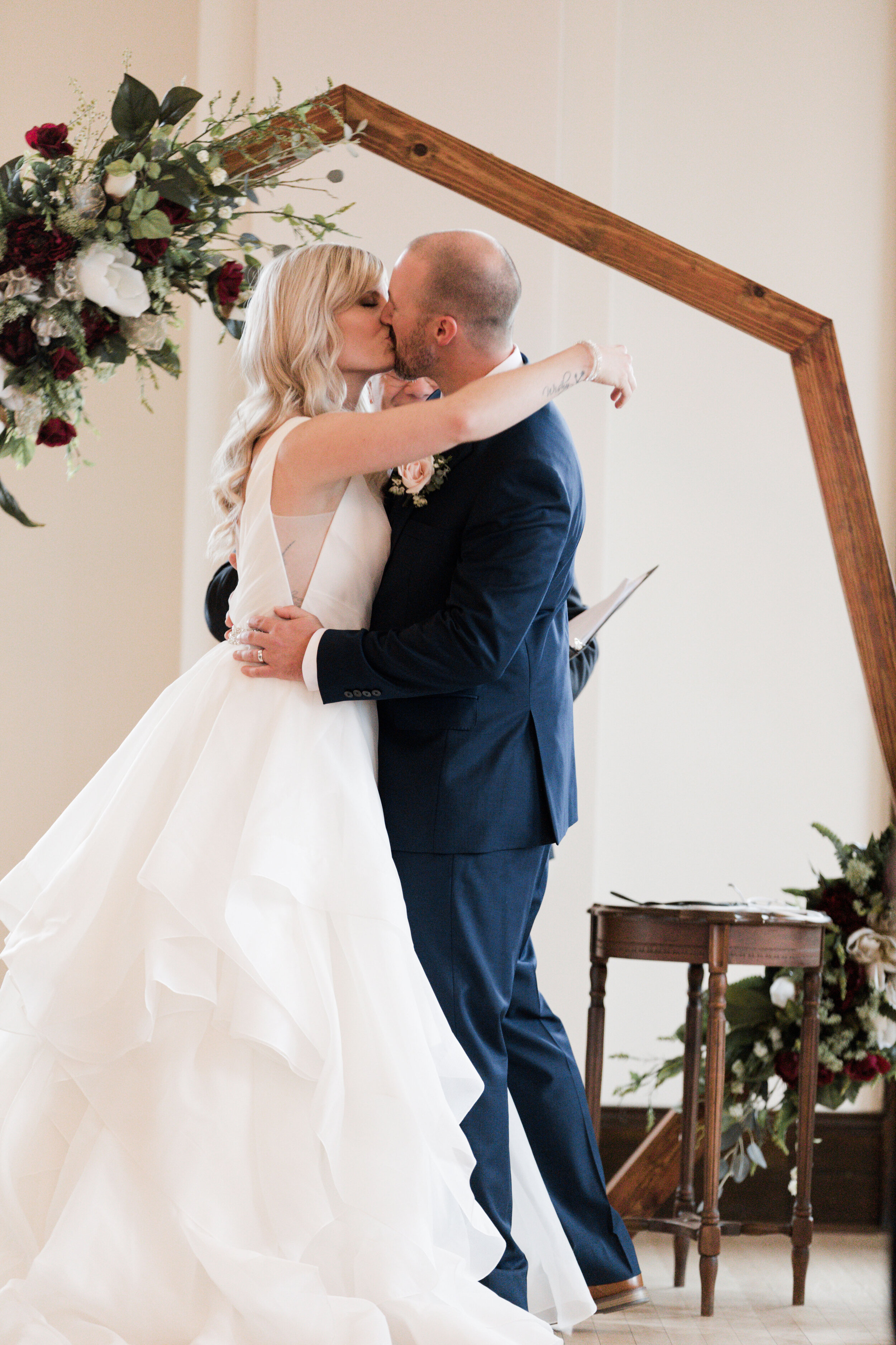 A bride and groom kissing during their wedding ceremony, standing under a wooden arch decorated with a floral arrangement of white, burgundy, and green flowers.