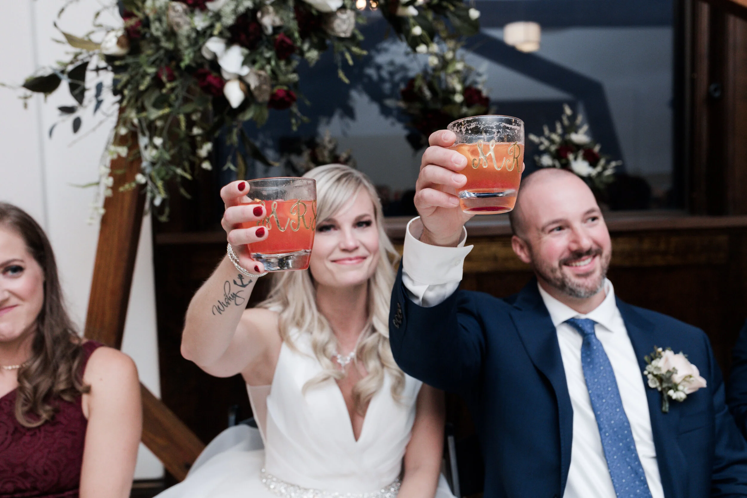 A wedding toast with a bride and groom holding glasses of rosé wine. The bride is smiling at the camera with a white dress, and the groom is also smiling, wearing a dark suit with a boutonnière. In the background is a floral arrangement with white an