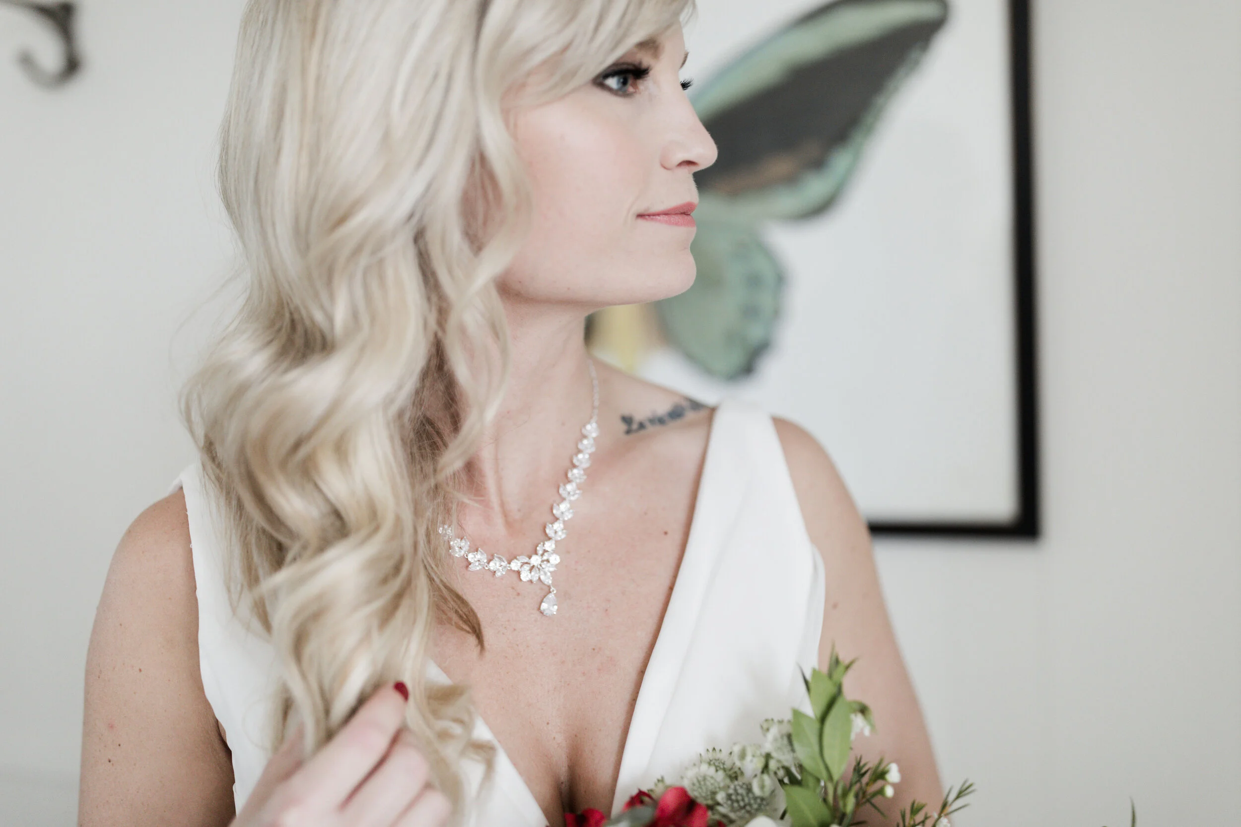 A woman with long blonde hair in waves, wearing a white dress, a diamond necklace, and holding a bouquet of flowers, looking to the side.