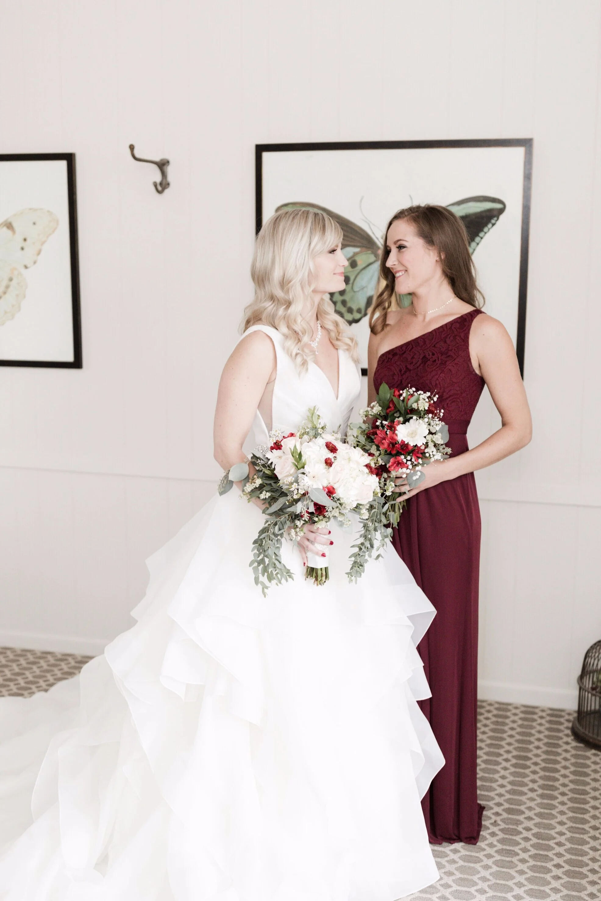 Two women, one in a white wedding gown with a bouquet, and the other in a burgundy dress, standing close and smiling at each other in front of framed butterfly pictures on a white wall.
