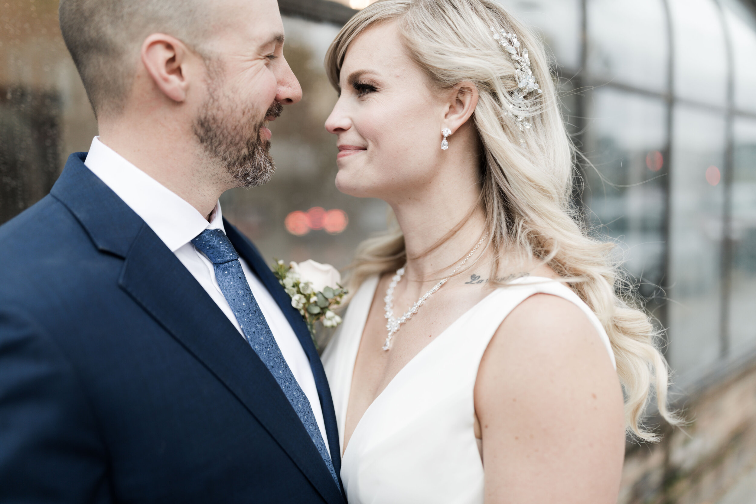 A bride and groom face each other closely, smiling, during their wedding. The groom wears a dark suit, white shirt, and blue tie, with a white boutonnière on his lapel. The bride has long blonde hair, adorned with a decorative hairpiece, wearing a wh