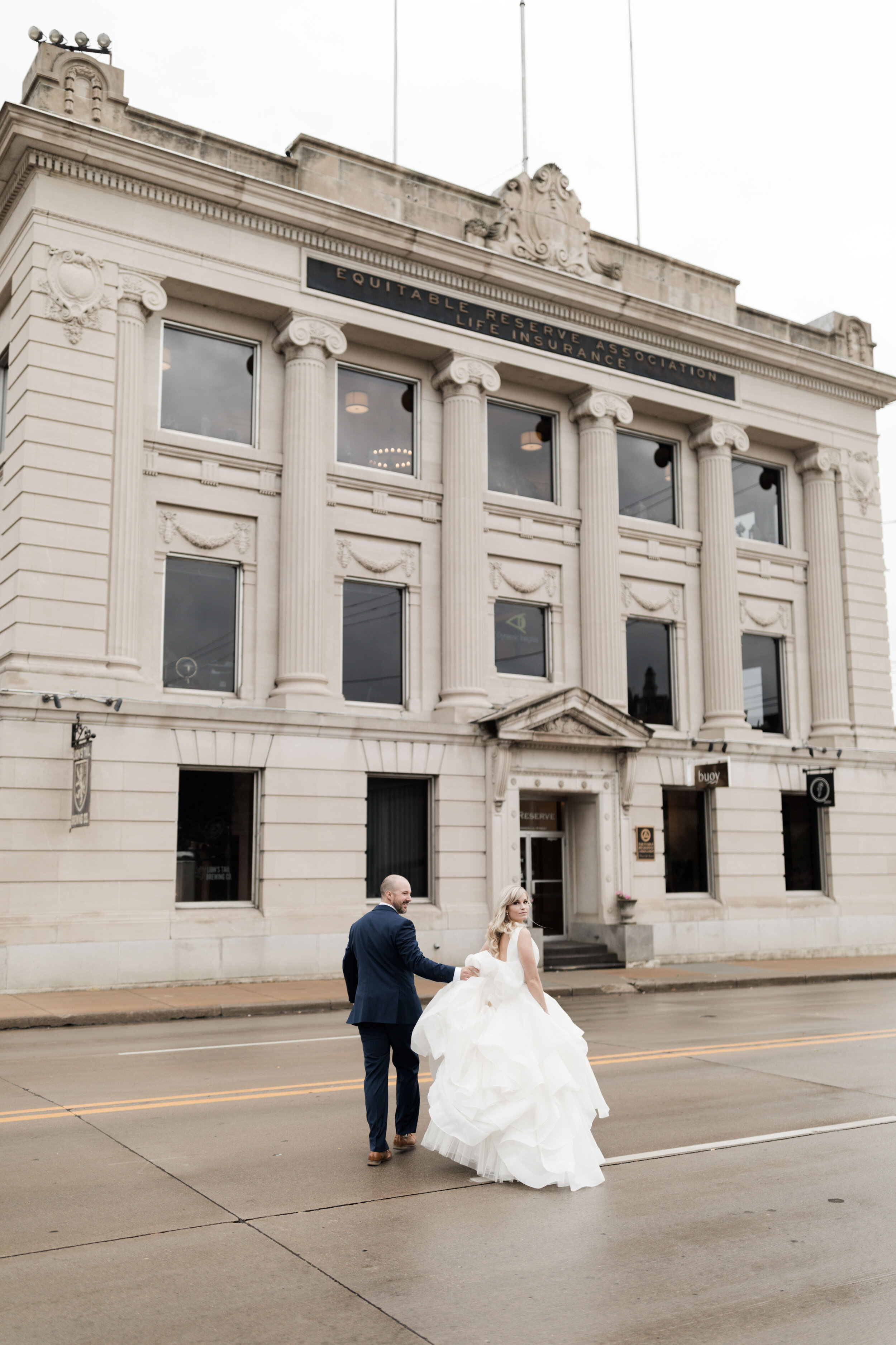 A newlywed couple walking on the street in front of a historic building. The bride is in a white wedding gown, and the groom in a navy suit. The building features classical architecture with columns and an inscription above.