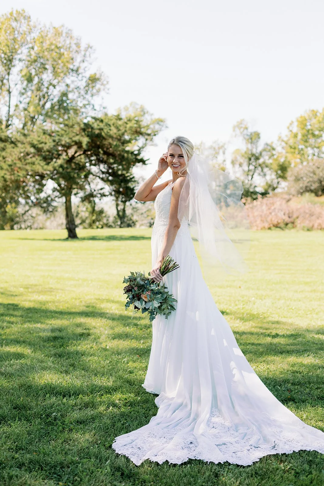 A bride in a white wedding dress holding a bouquet of greenery and flowers, standing outdoors on a green lawn with trees in the background, smiling.