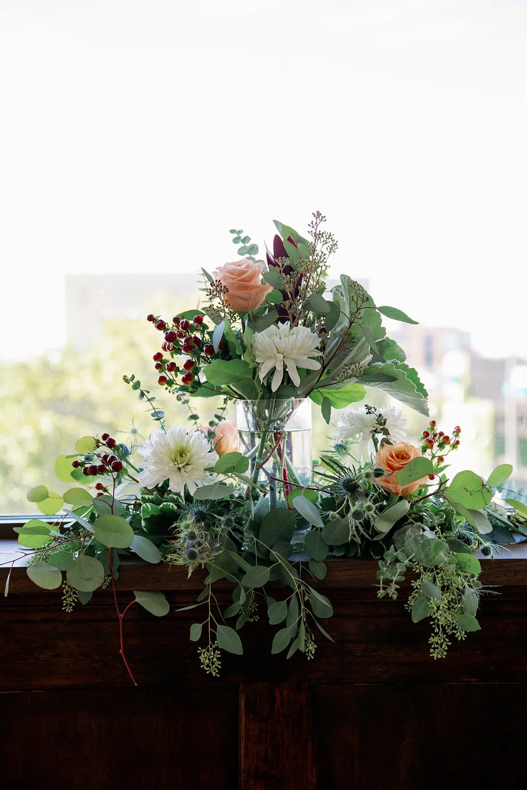 A floral arrangement in a glass vase on a wooden surface near a window, featuring peach roses, white daisies, red berries, and green foliage.
