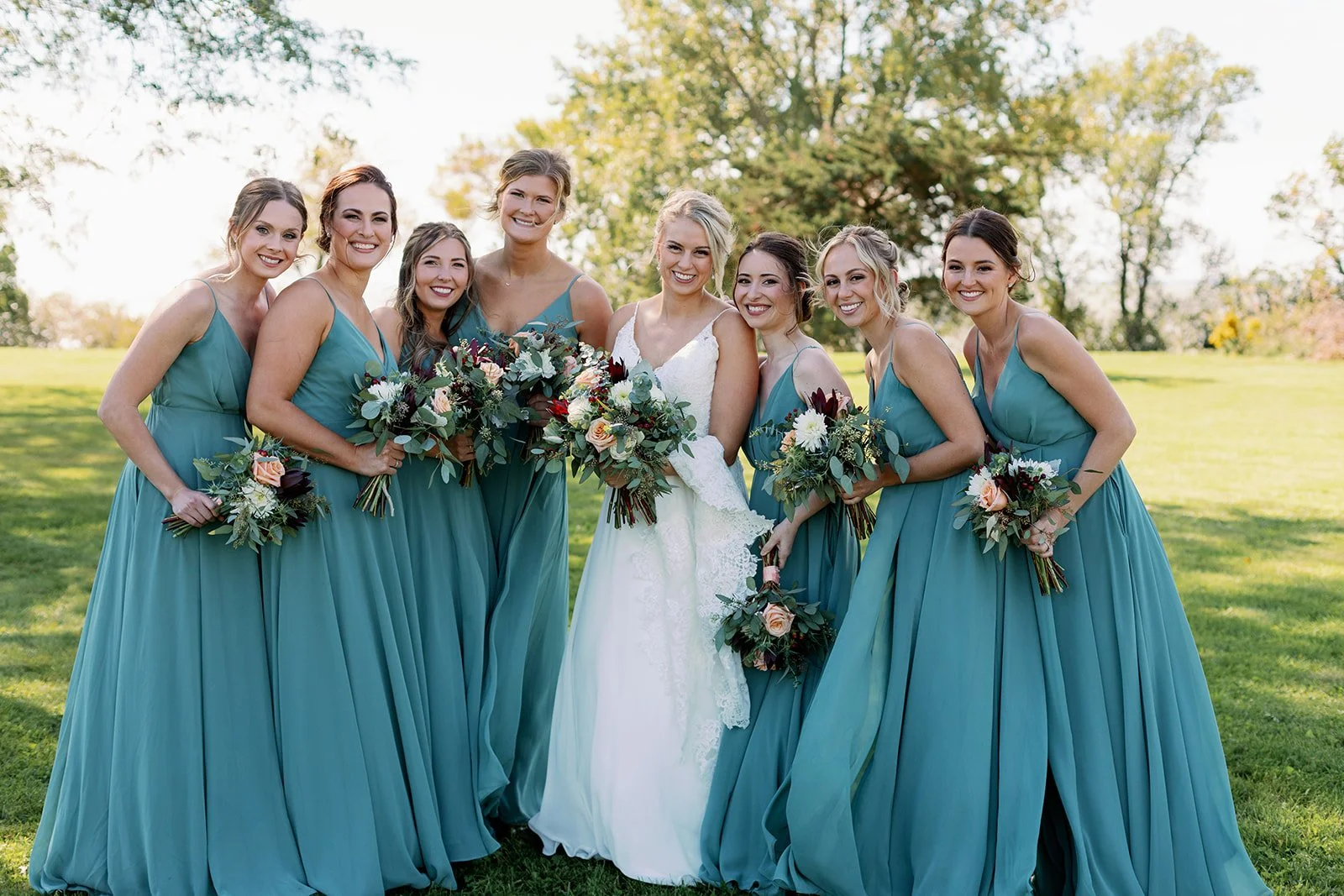 Group of women, including the bride in a white wedding dress, standing outdoors on a grassy field with trees in the background, all smiling and holding bouquets, during a wedding celebration.