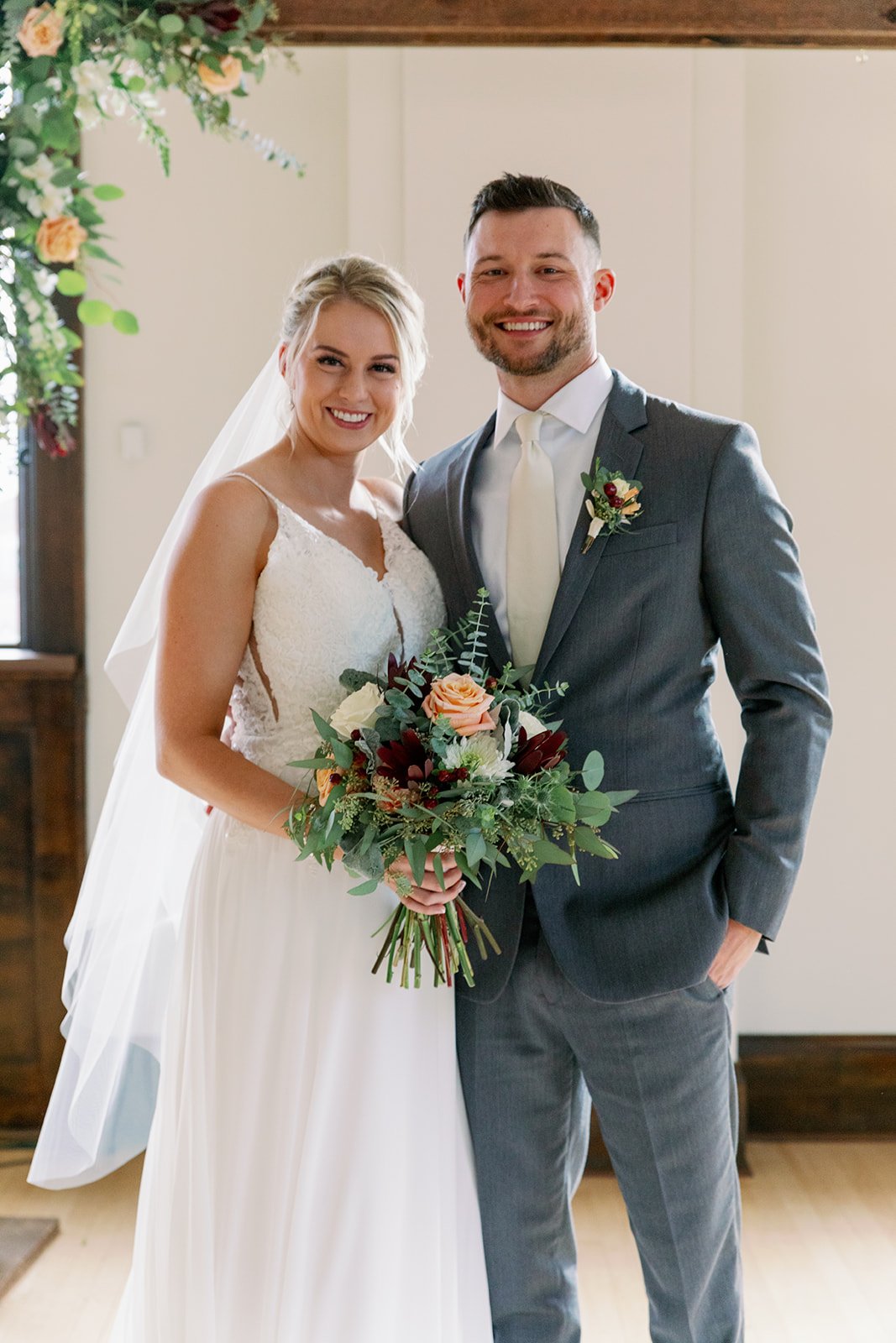 A bride and groom on their wedding day, standing indoors and smiling at the camera. The bride is holding a bouquet of flowers, and the groom is dressed in a gray suit with a boutonniere.