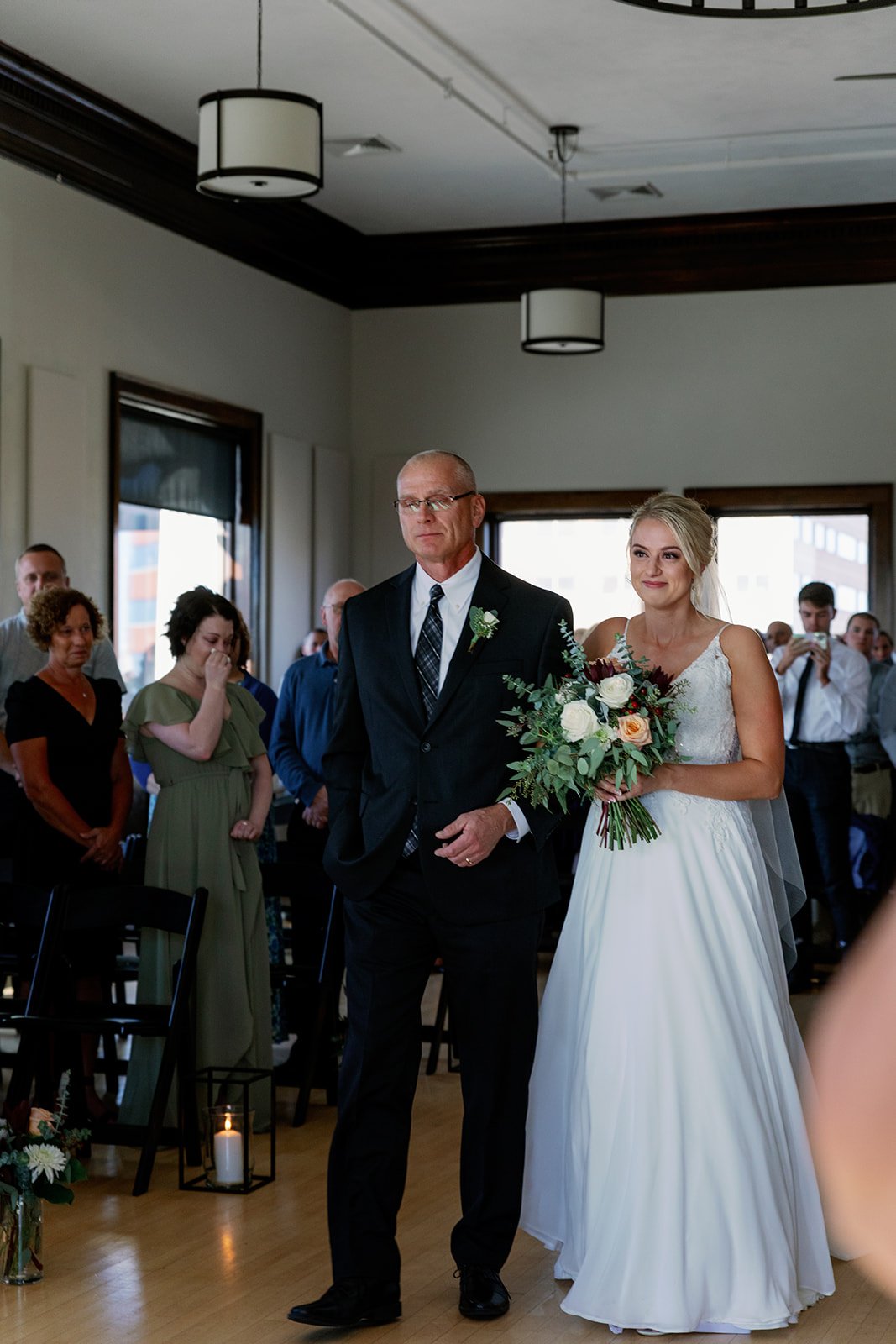 A bride in a white wedding dress holding a bouquet of flowers walking down an aisle with her father during a wedding ceremony. People are standing and watching in the background.