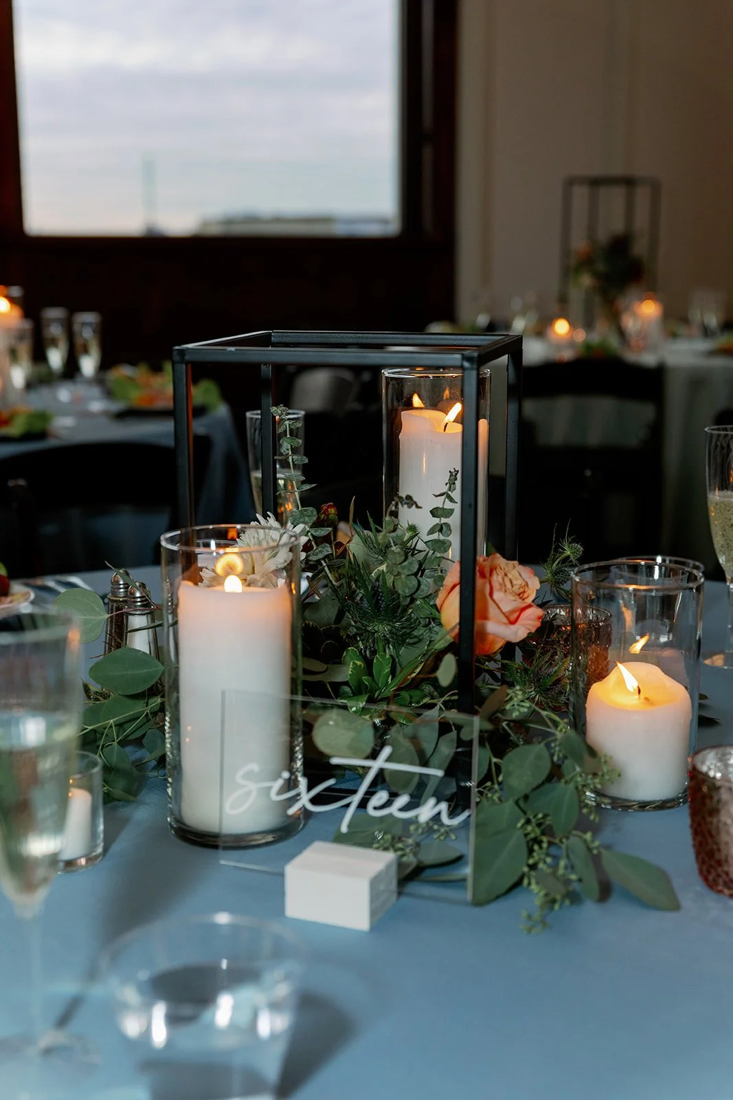 Elegant wedding reception table centerpiece with lit candles, greenery, and a glass sign reading 'sixteen'.