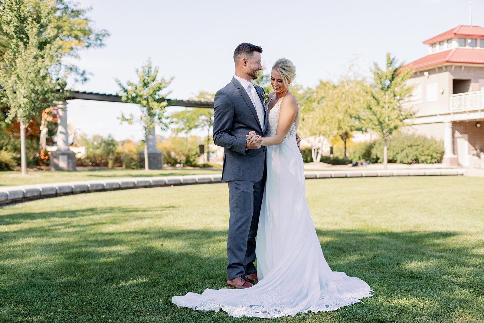A bride and groom share a dance outdoors on a sunny day, with trees and a building in the background.