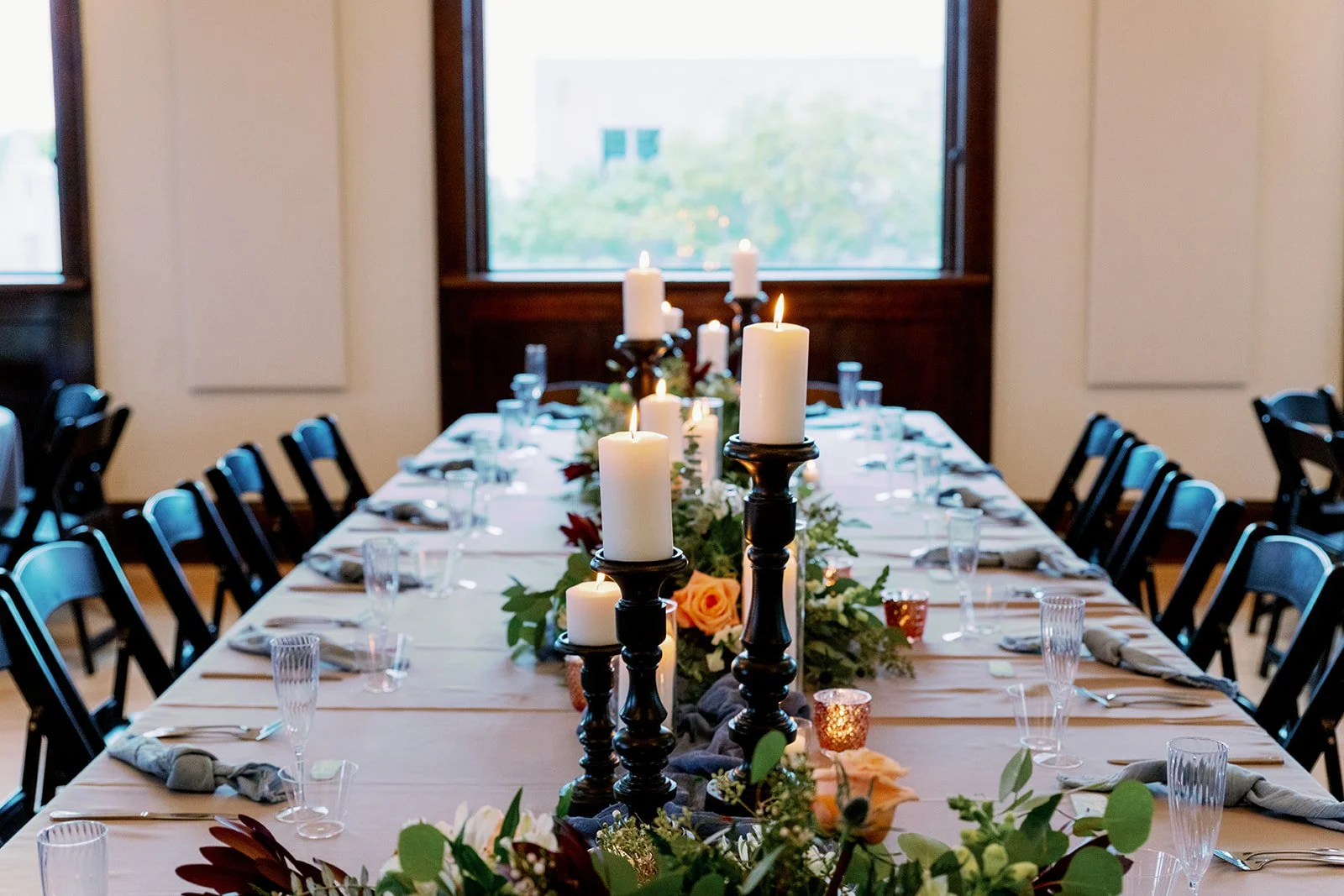 A long dining table decorated with candle centerpieces, flowers, and tableware, set for a formal event with chairs around it, in a room with large windows.