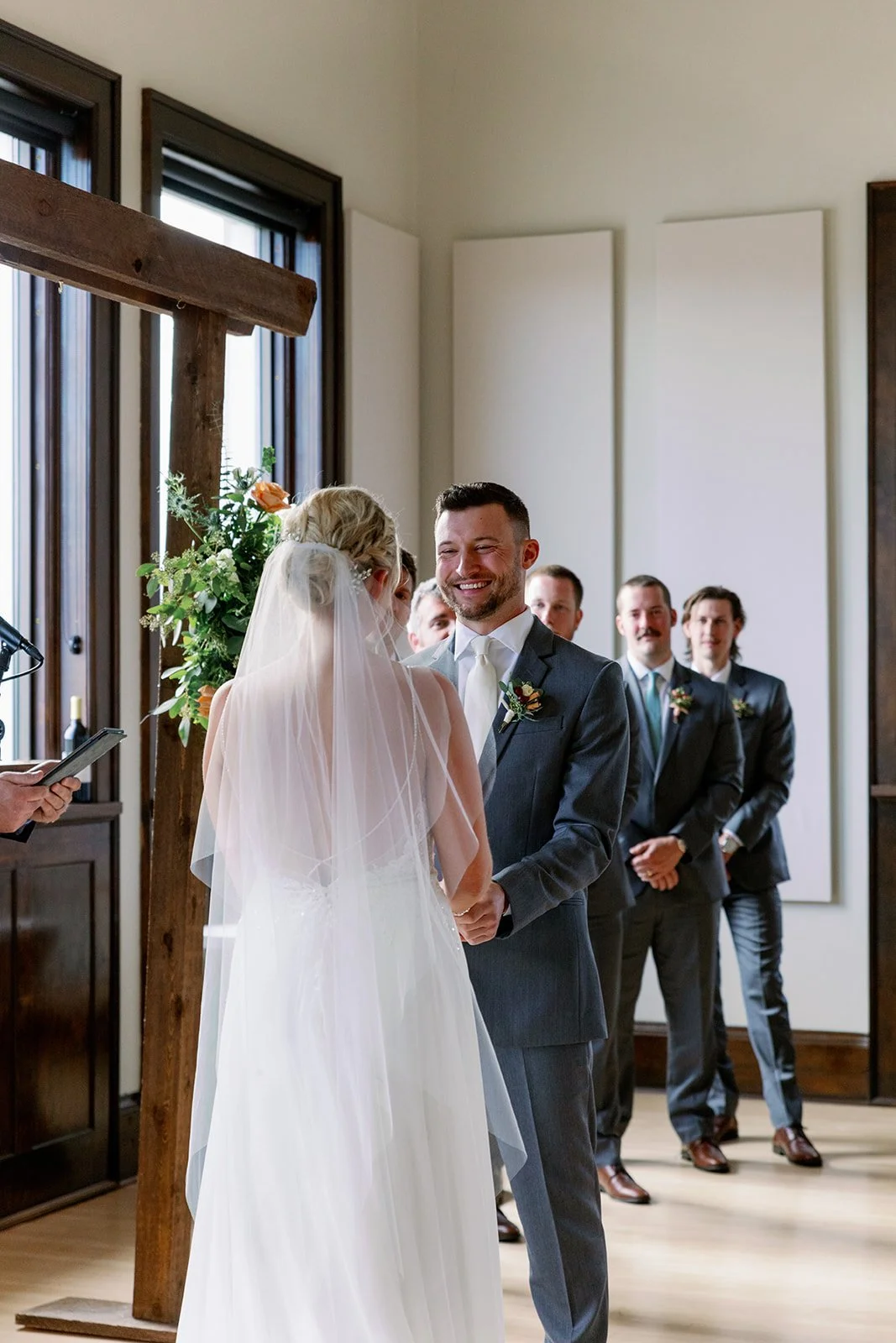 A bride and groom exchange vows during a wedding ceremony indoors, with groomsmen standing behind them, a wooden cross, and floral arrangements.