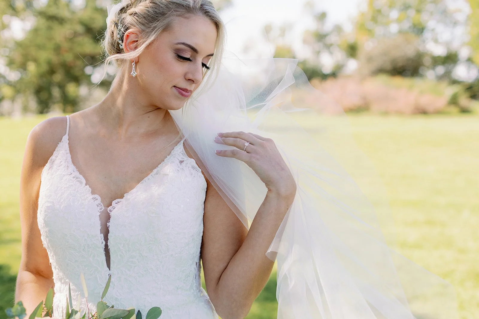 Bride in a white lace wedding dress with a tulle veil outdoors holding a bouquet.