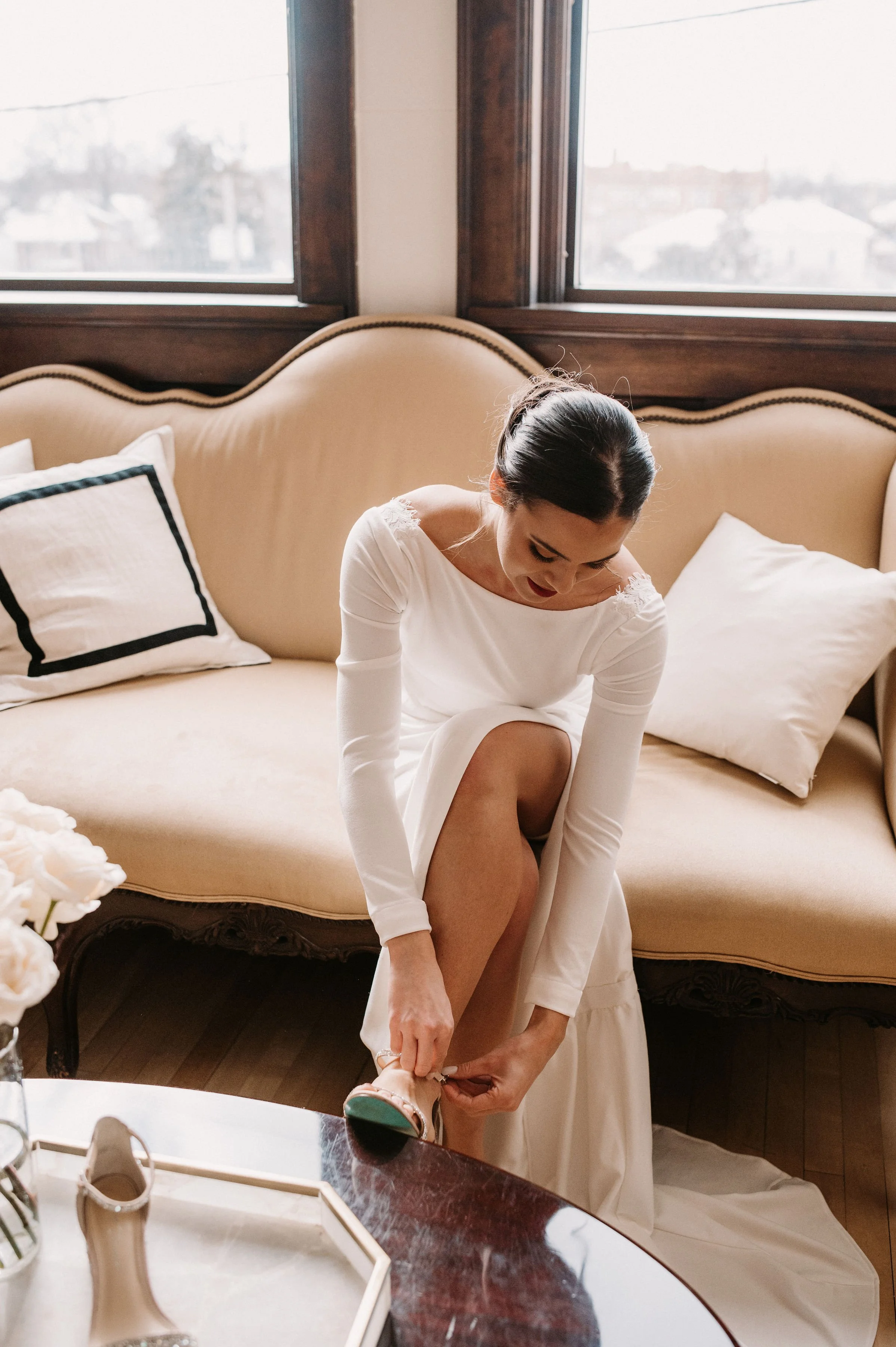 Woman in white dress putting on her shoe in a living room with a beige vintage-style couch and large windows.