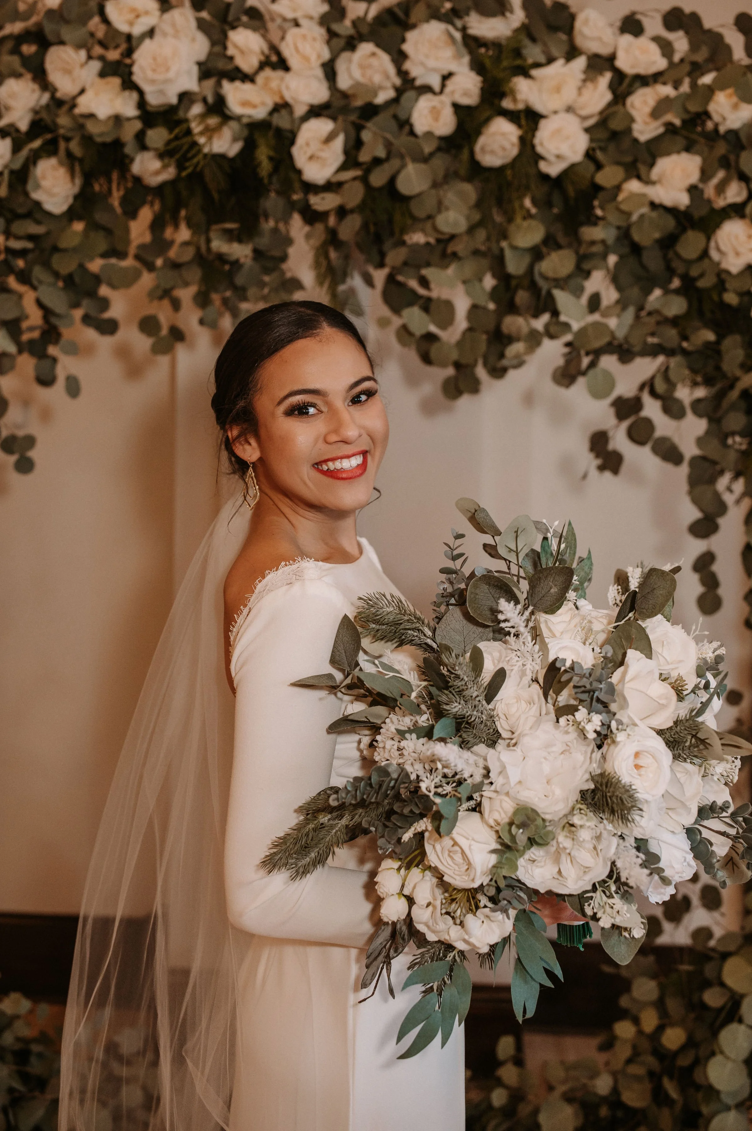 A smiling bride holding a large bouquet of white roses, eucalyptus, and greenery, standing in front of a floral backdrop with white roses and eucalyptus.