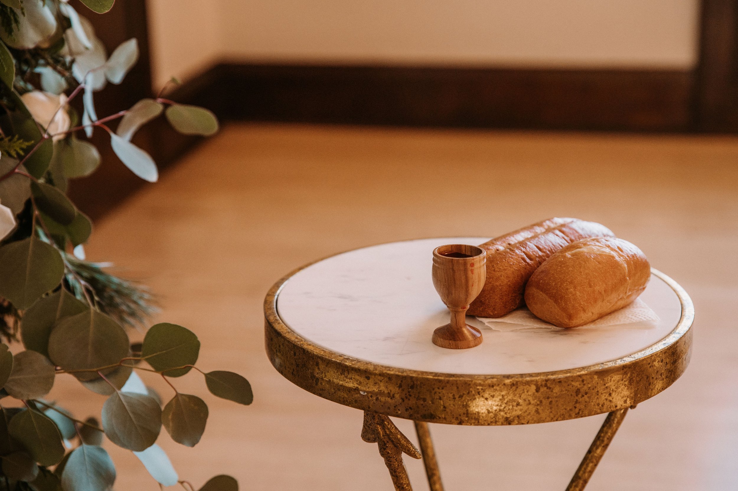 A round table with a rustic metal edge holds two loaves of bread and a small wooden goblet, with green leaves on the left side.