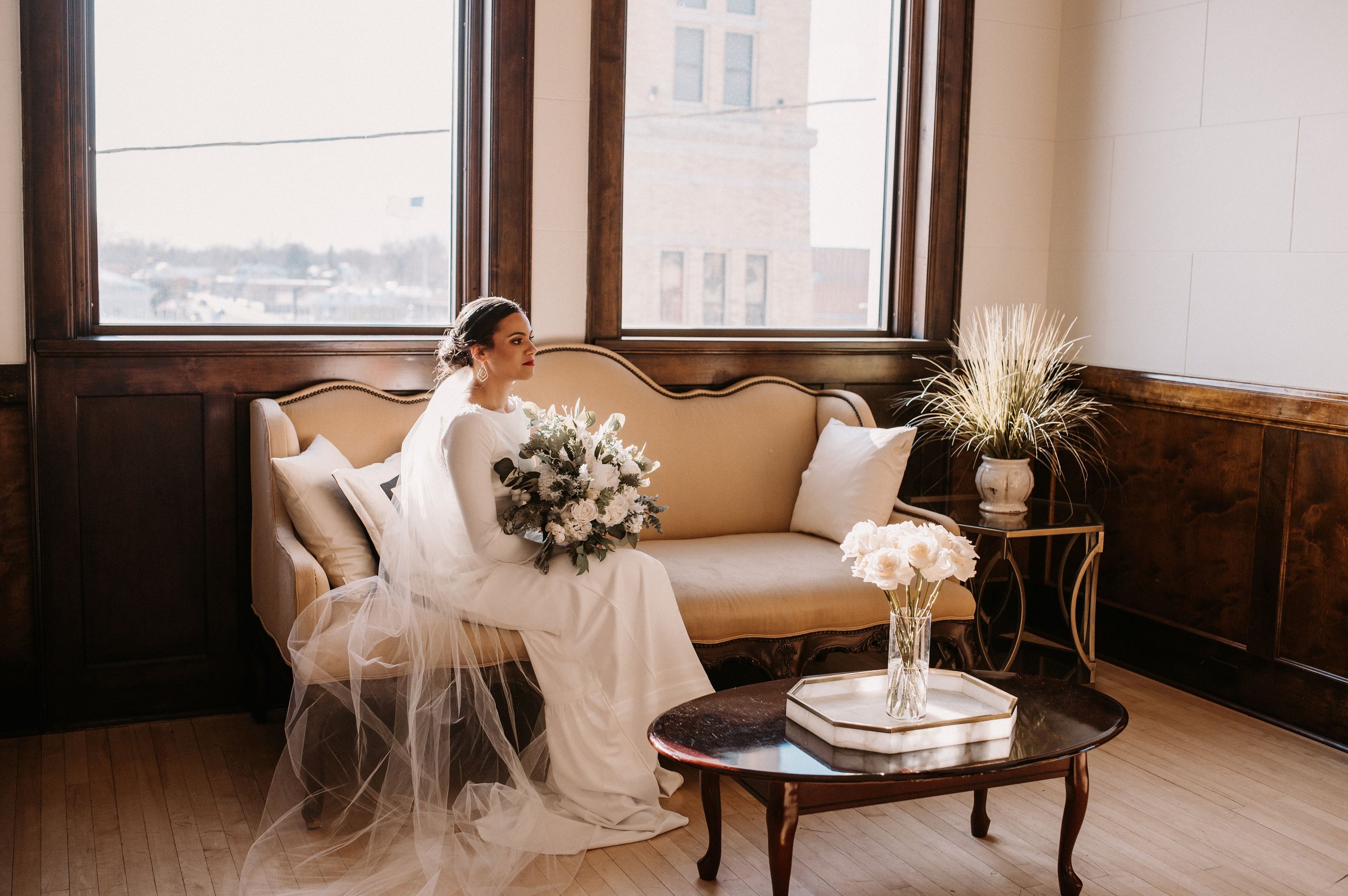 Brunette bride in a white wedding gown and veil sitting on a beige vintage sofa, holding a bouquet of white flowers, in a sunlit room with large windows, wooden trim, white walls, and decorative plants.
