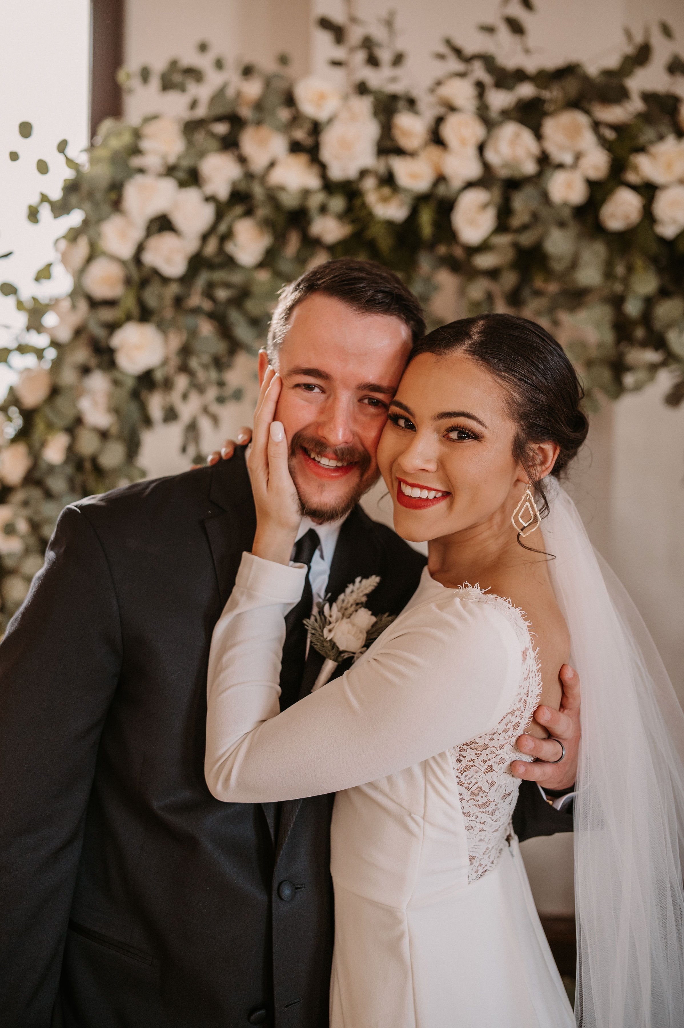 A bride and groom pose closely during their wedding celebration, smiling with a floral backdrop behind them.