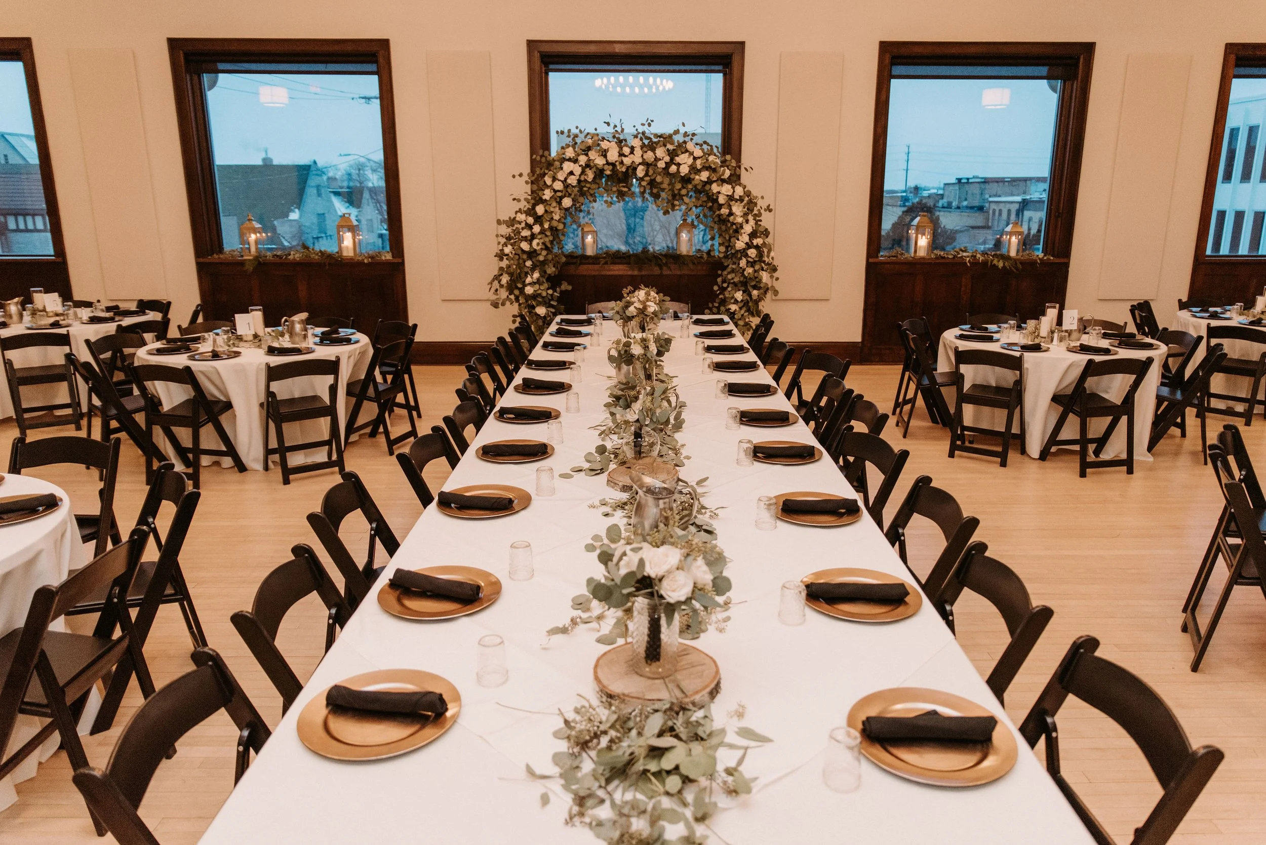Wedding reception table setup with a long white table, centerpieces of greenery and white flowers, and a floral arch in the background along with windows and lantern decorations.