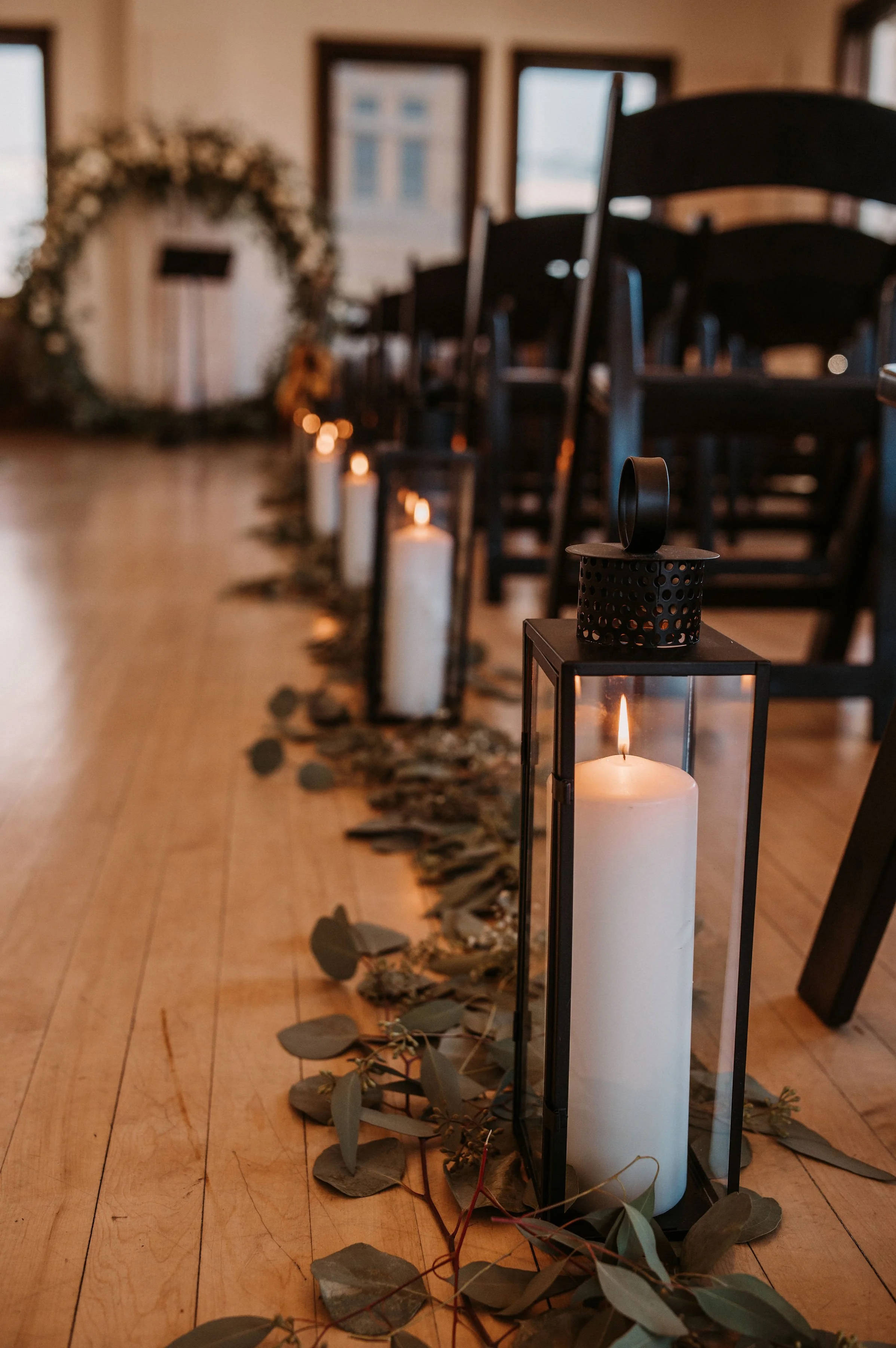 Lit candles inside glass lanterns arranged in a line on the floor, with eucalyptus leaves scattered around, in a decorated indoor event space with chairs and a floral arch in the background.