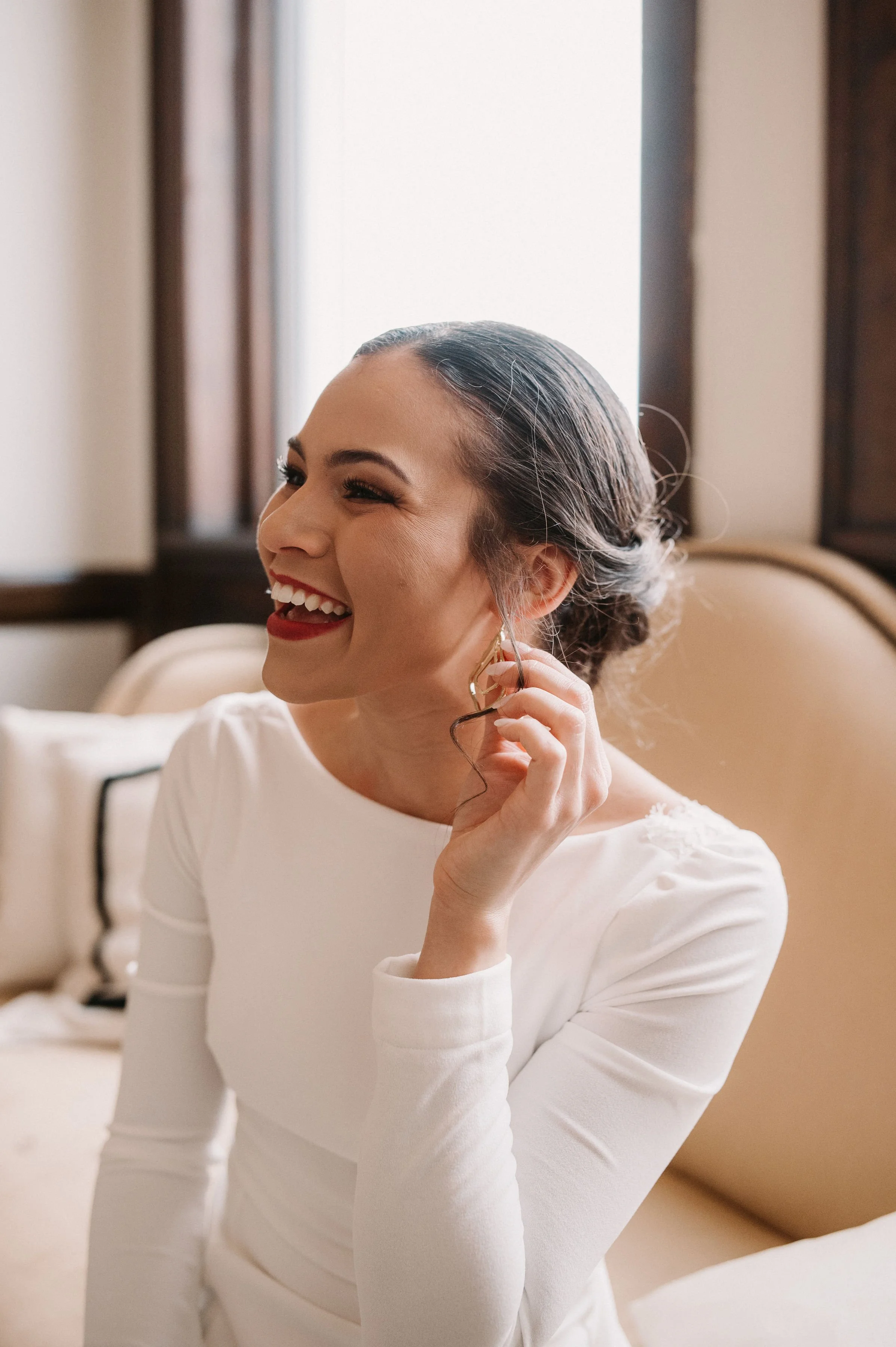 Woman smiling and adjusting her earring indoors, wearing a white dress with dark hair styled in an updo.