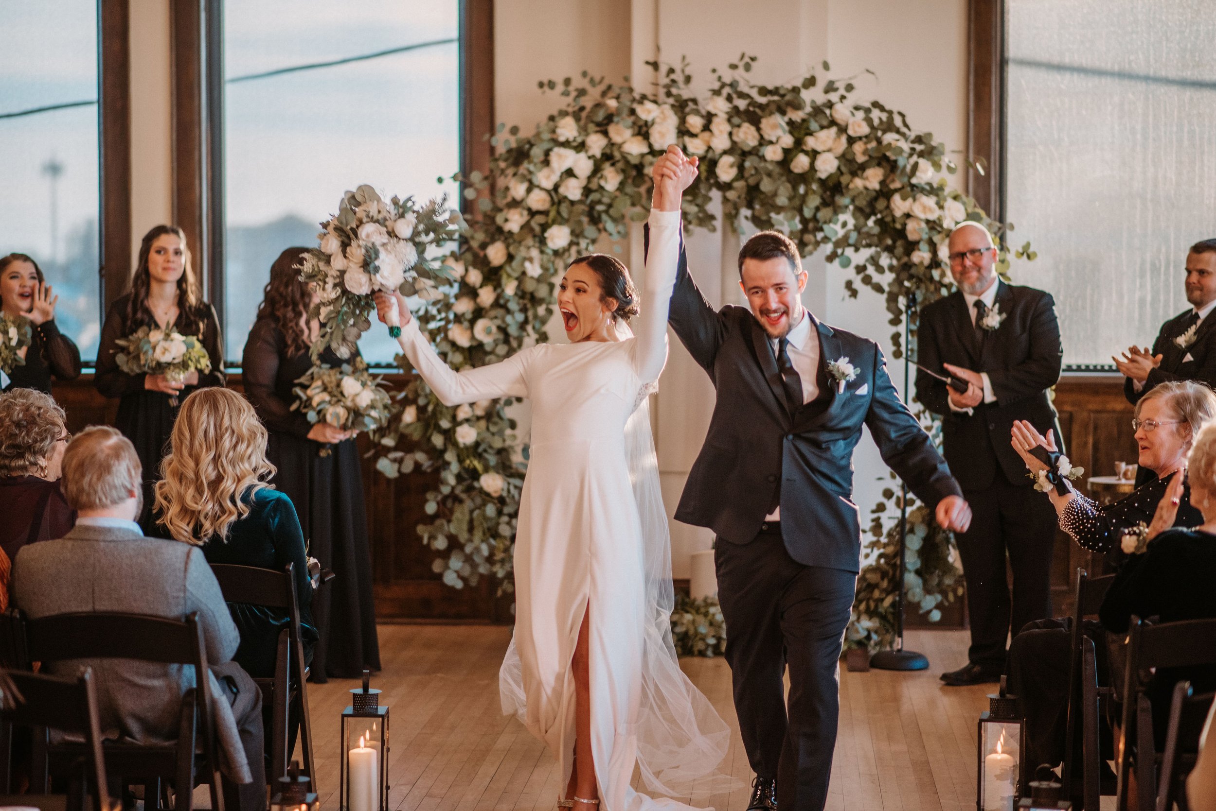 Bride and groom celebrate their wedding while walking down the aisle, holding hands and raising arms, surrounded by cheering guests and floral decorations.