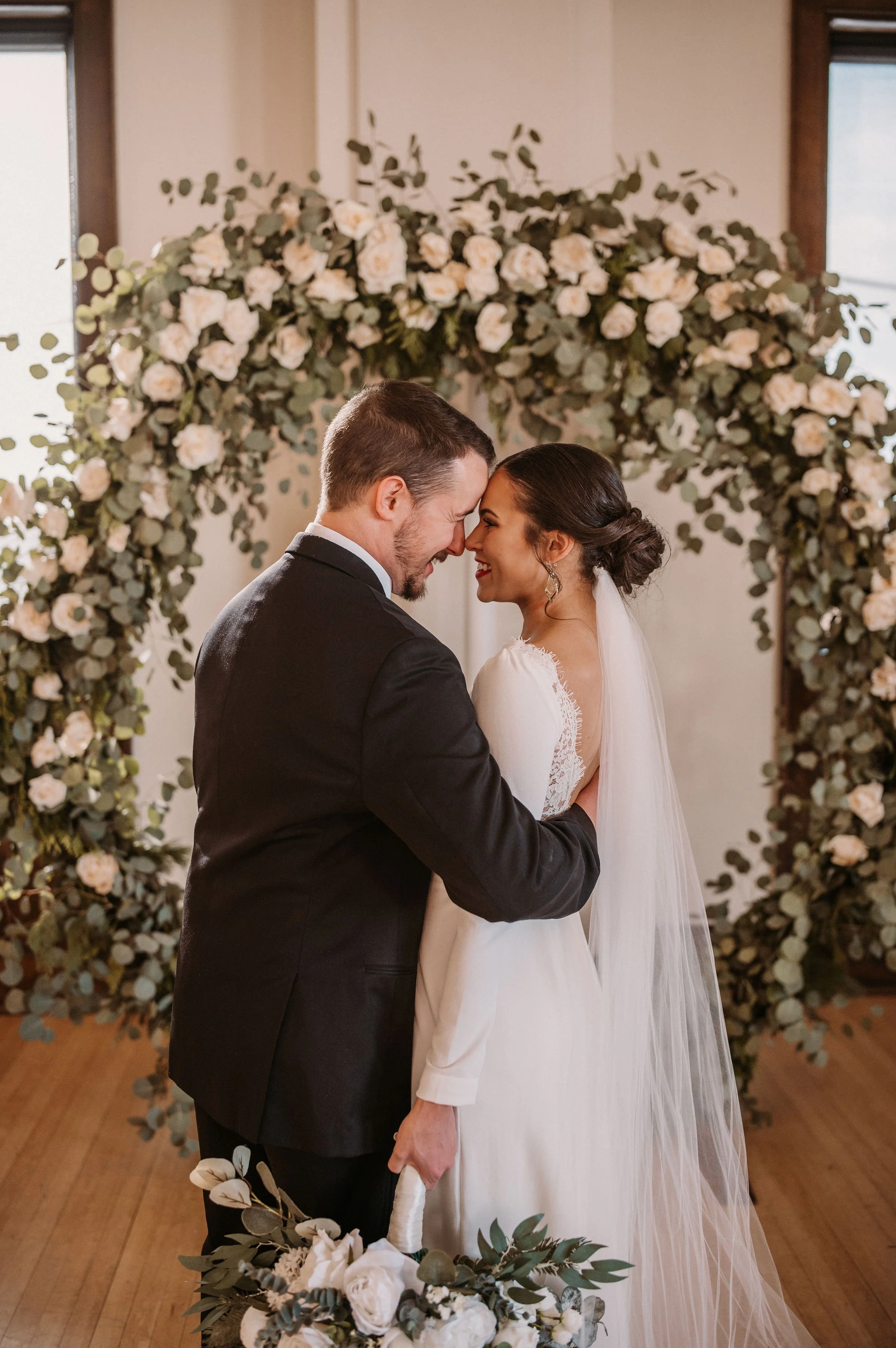 A bride and groom sharing a kiss at their wedding ceremony, standing in front of a floral arch decorated with white roses and greenery.