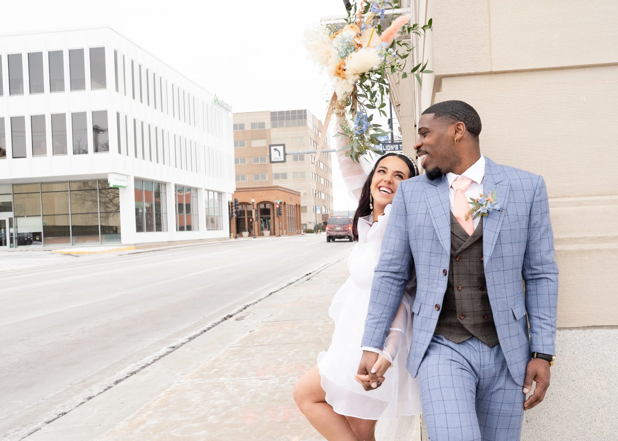 A smiling couple walking hand in hand on a city sidewalk, with the woman holding a bouquet of flowers over her head, both dressed in wedding attire.