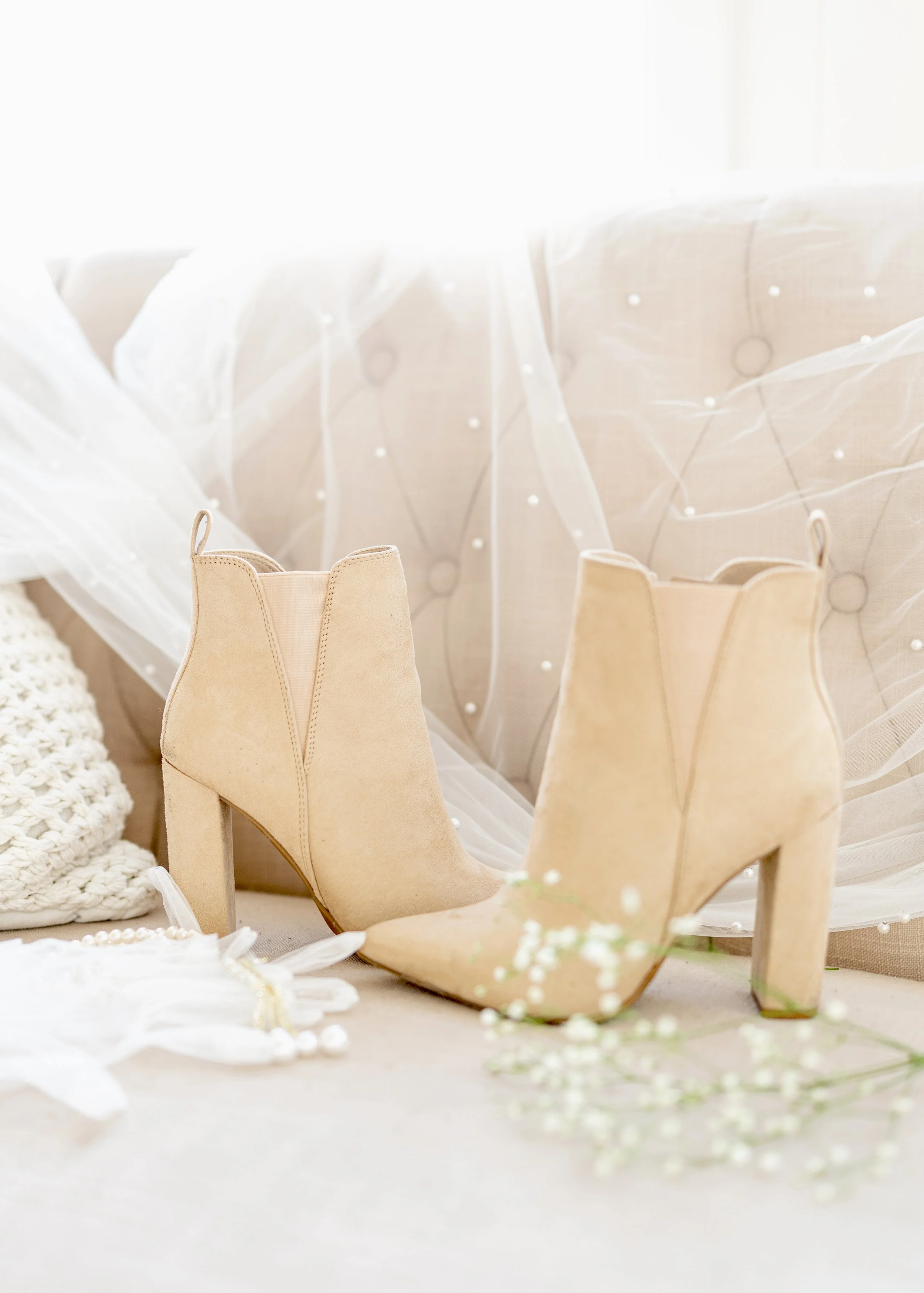 A pair of beige high-heeled ankle boots placed on a light-colored surface, with a soft, vintage background that includes a crocheted item and decorative tulle fabric with small white pearls.