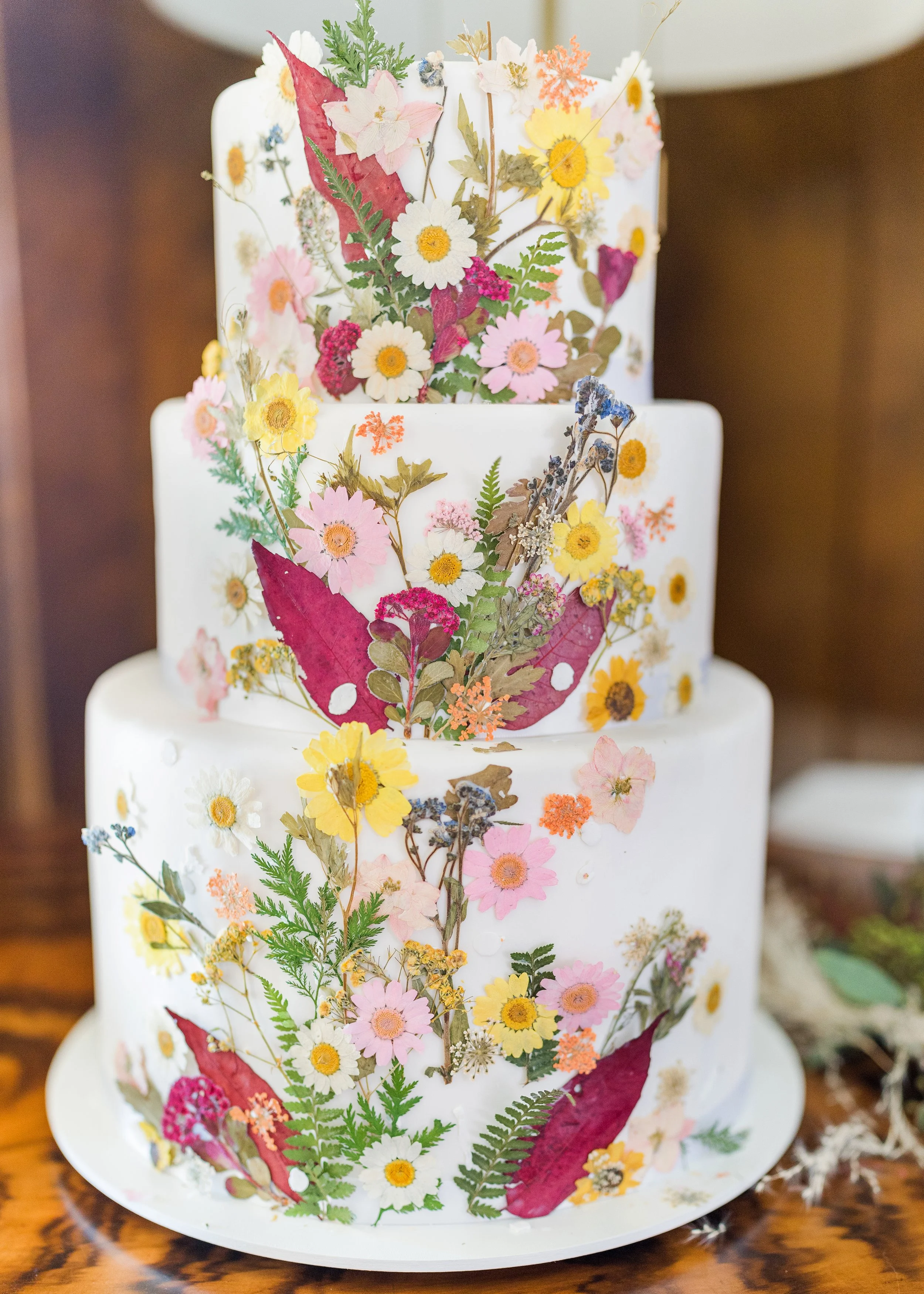 Three-tiered white wedding cake decorated with colorful edible flowers and leaves, including pink, yellow, purple, and red blooms, along with green fern leaves, arranged in a natural, botanical style.