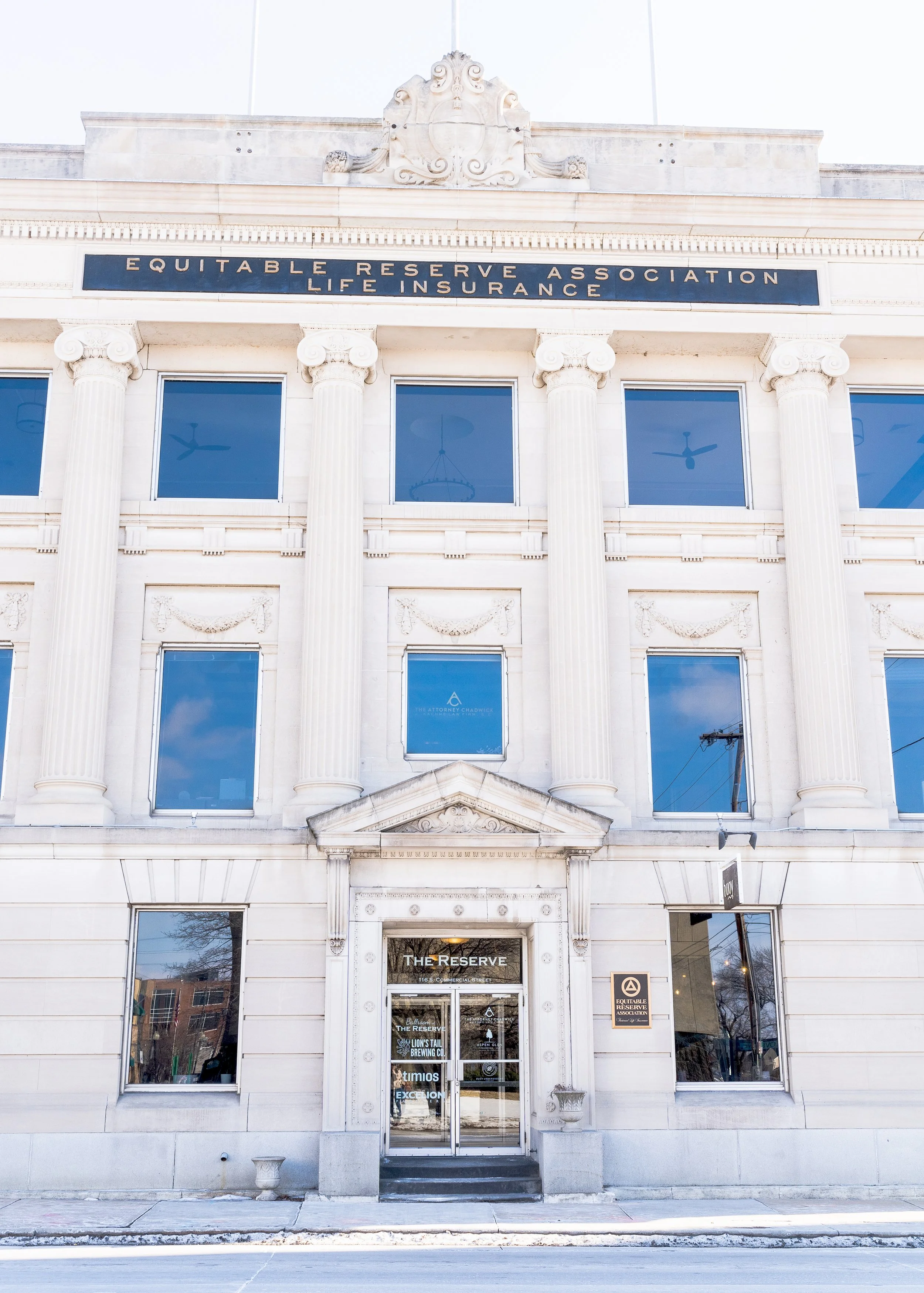 Front view of a white, classical-style building with four columns and an ornate pediment. A sign on the building reads 'Equitable Reserve Association Life Insurance.' The building has large windows and a glass door with the name 'The Reserve' above i