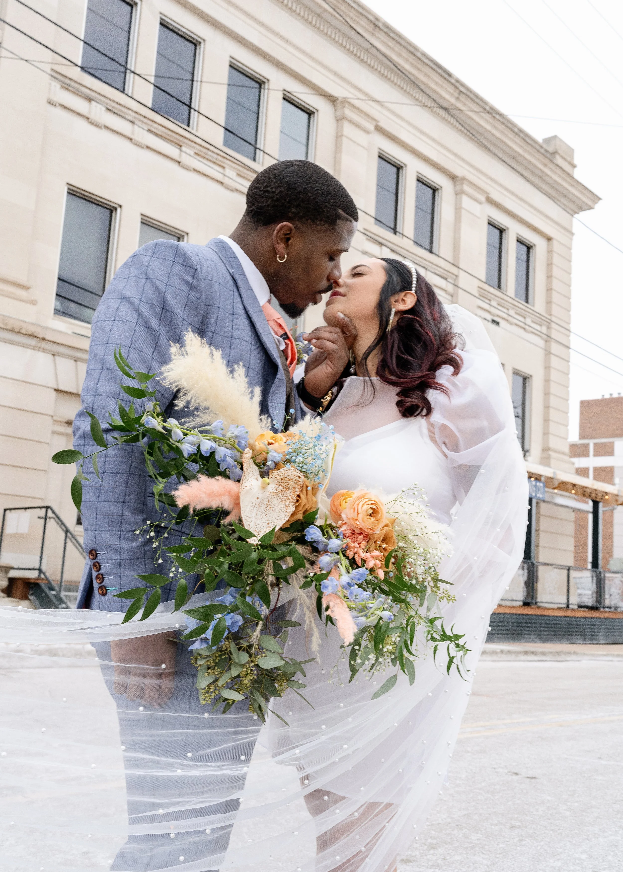 A couple dressed in wedding attire sharing an intimate moment outdoors, with the man in a blue checkered suit and the woman in a white dress holding a bouquet of colorful flowers.