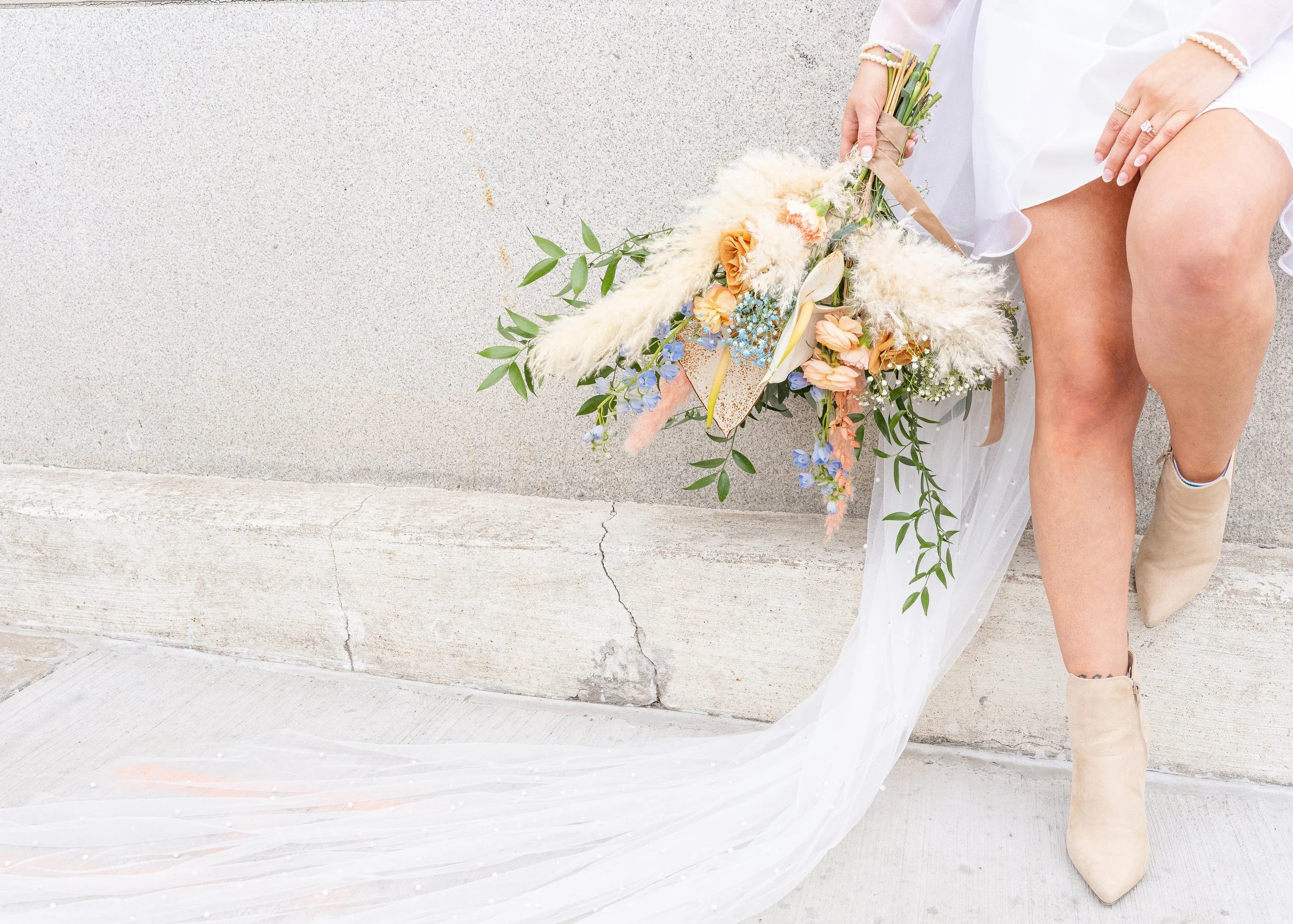 A person in a white dress and beige boots holding a large bouquet of flowers, standing against a concrete wall.