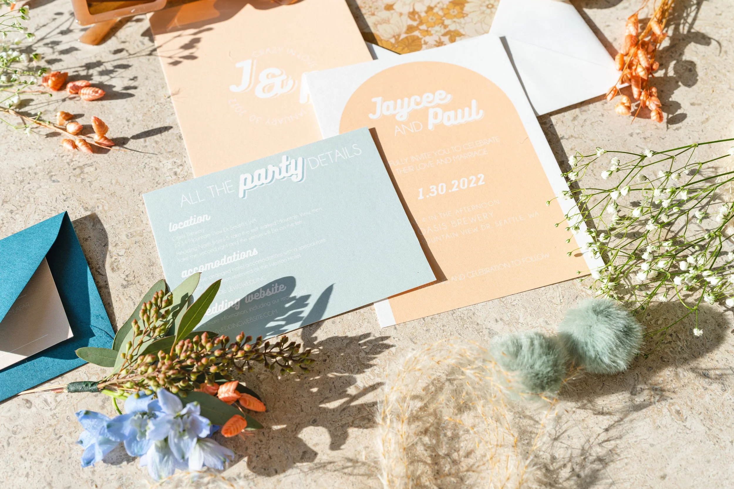 Colorful wedding invitation cards, floral decorations, and an open blue envelope arranged on a sandy surface with shadows of flowers and greenery.