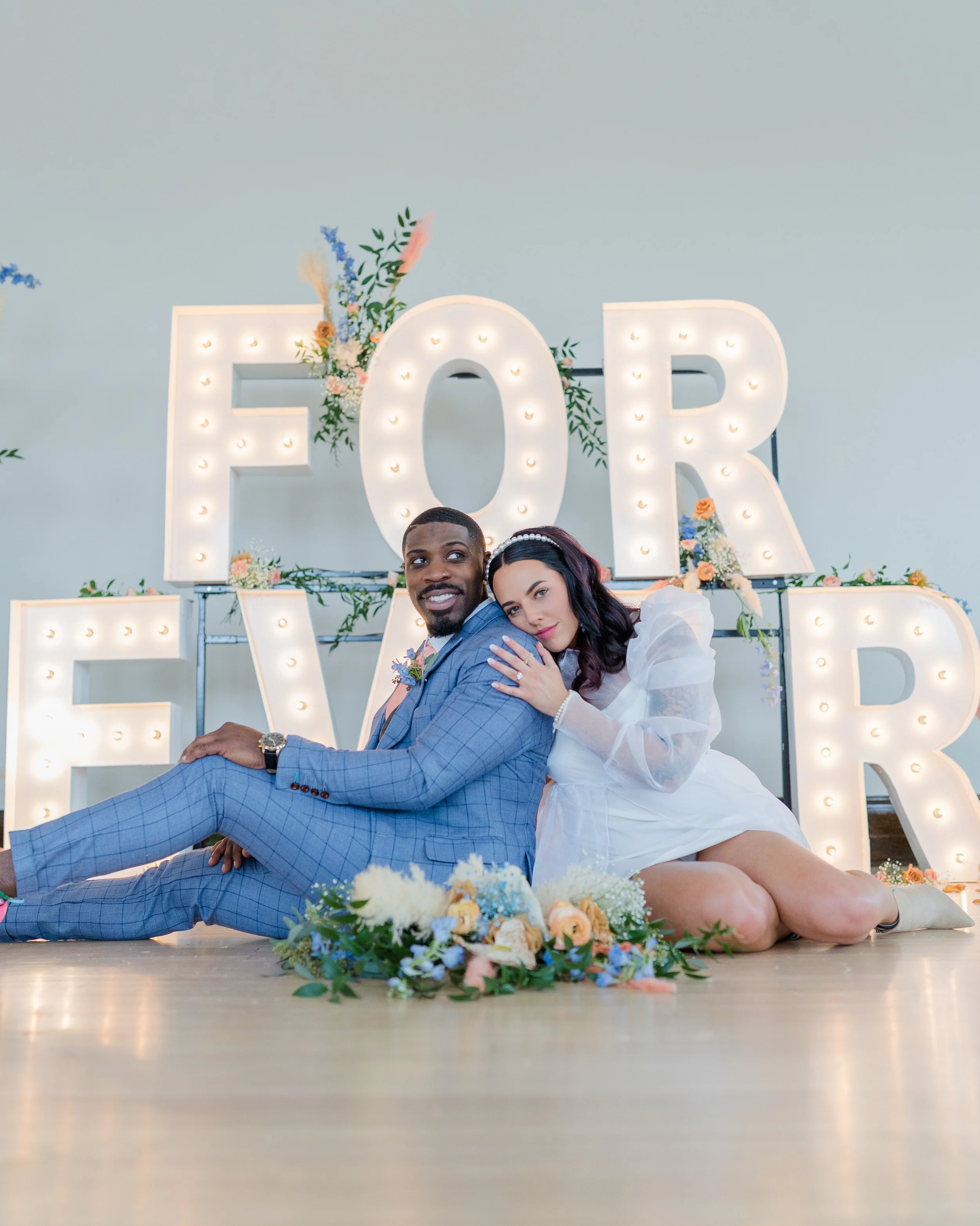 Happy couple sitting on the floor in front of large illuminated "For Ever" sign decorated with flowers at their wedding celebration.