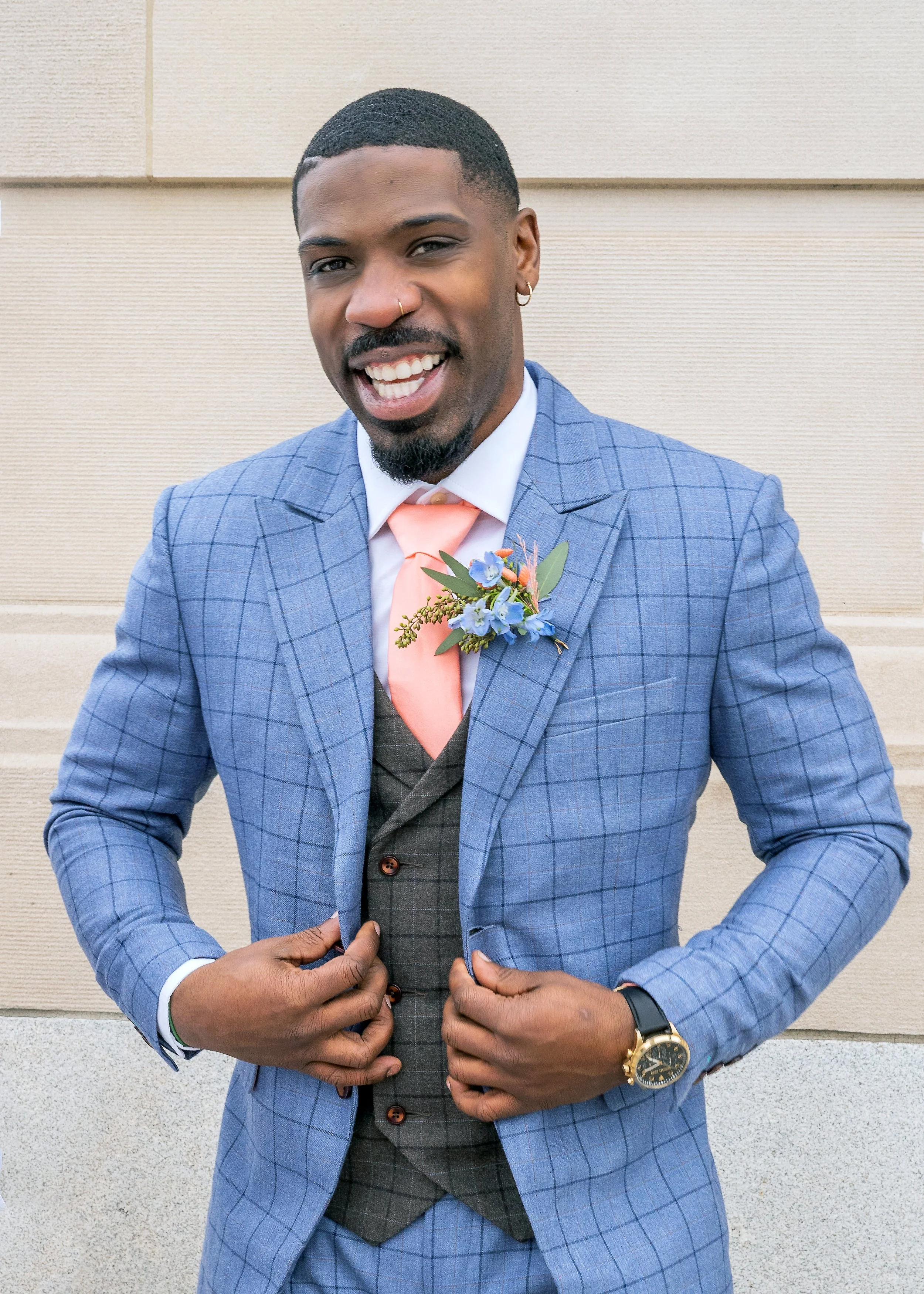 A man wearing a blue checkered suit, pink tie, gray vest, and boutonniere with blue flowers, smiling and adjusting his jacket, standing against a beige wall.