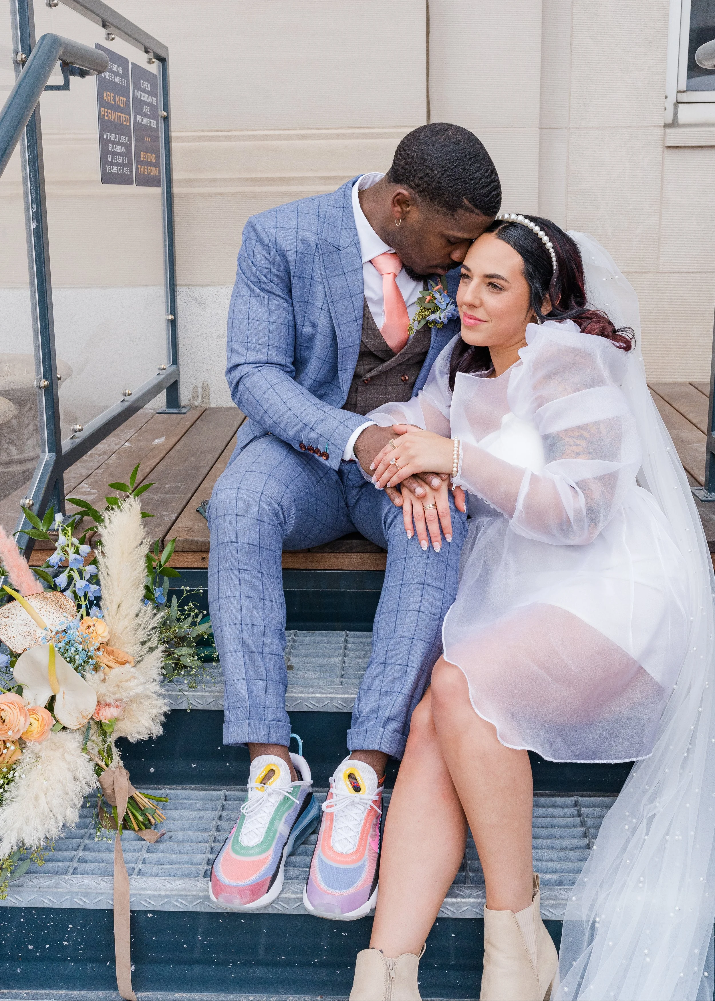 A bride and groom sitting on steps outside, with the groom gently kissing the bride's forehead. The bride is wearing a white wedding dress and veil, and the groom is in a light blue checkered suit with sneakers. There is a floral bouquet of peach, wh
