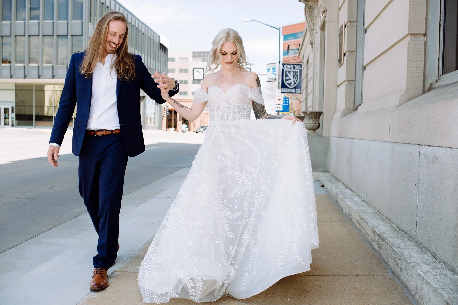 A couple, with the woman in a wedding dress and the man in a suit, walking on a city sidewalk and holding hands.